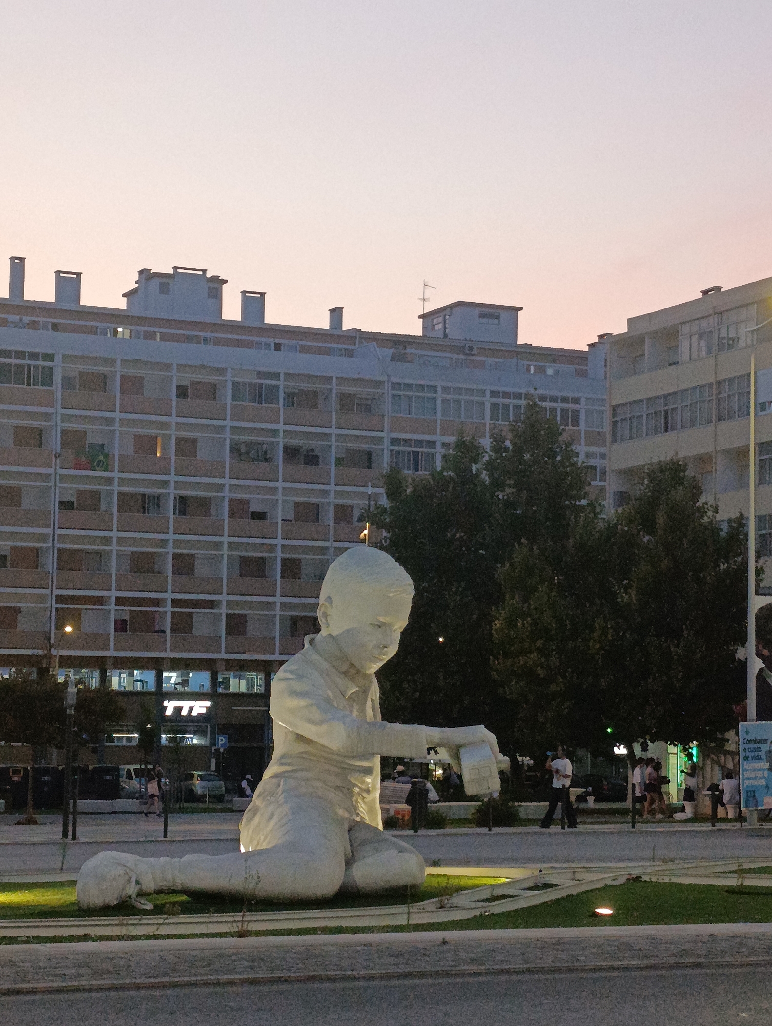 The photograph captures a large, white, chalk-like sculpture of a child in a seated position, leaning forward with one arm extended. The sculpture is located in a public plaza in Setúbal, Portugal, likely in front of the buildings that line the plaza. The background features a multi-story apartment building with numerous balconies and windows, typical of urban residential architecture. Trees are visible behind the sculpture, and in the distance, other buildings and streetlights can be observed. The sky above is a soft, pale orange, indicating either dawn or dusk. Several people are present in the plaza, some walking, others conversing or sitting. The overall scene suggests a peaceful, urban environment during twilight hours. The sculpture itself is the primary focal point, rendered in a way that emphasizes its childlike form and its interaction with the surrounding space.