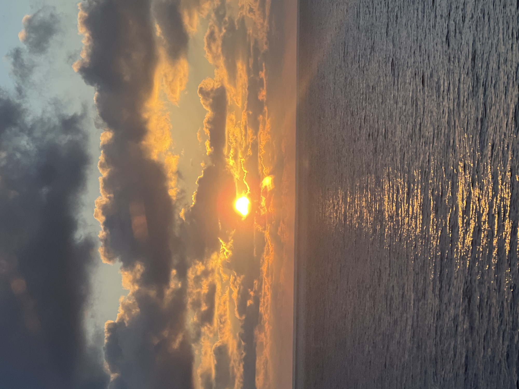 An image depicts a sunset over the ocean. The sun, a bright orb of orange and yellow, is partially obscured by clouds. These clouds are a mix of dark gray and illuminated with golden hues. The ocean below is calm, with a shimmering reflection of the sunset across its surface. The horizon line is clearly defined, separating the sky from the sea. The lighting suggests it is either sunrise or sunset, with warm tones dominating the scene. The location is unknown.