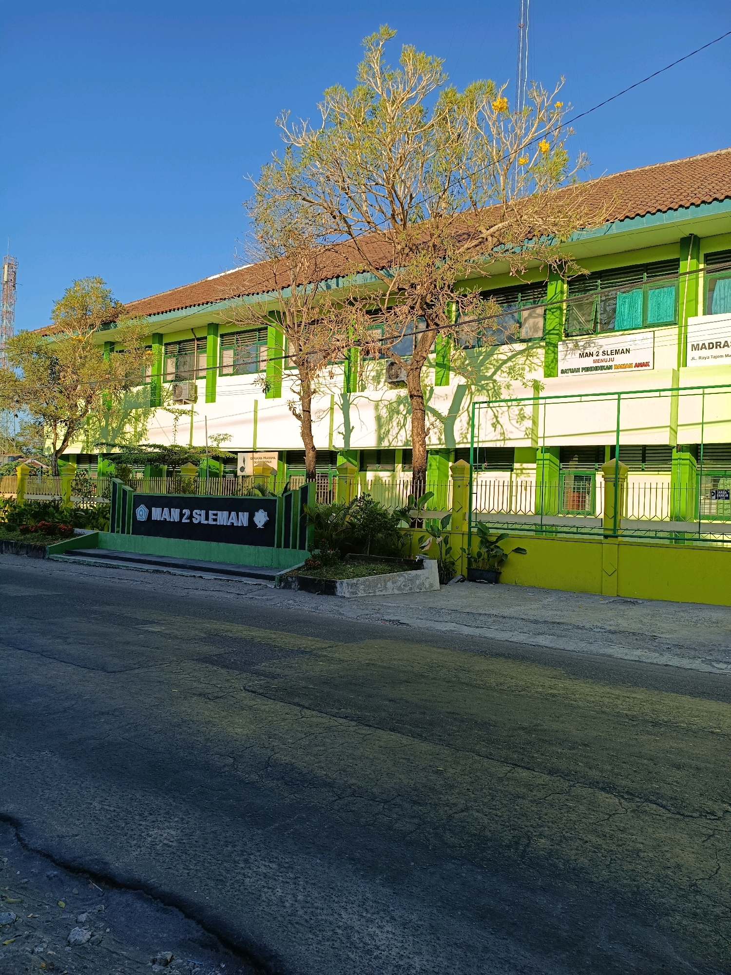 The image shows a two-story school building with a brown tiled roof and green and white walls, located in Maguwoharjo, Indonesia. A sign in front of the building reads "MAN 2 SLEMAN". There are trees in front of the building, casting shadows on the walls. The building appears to be an educational institution, possibly a Madrasah Aliyah Negeri (MAN), which is a public Islamic senior high school in Indonesia. The street in front of the school is paved with asphalt and shows some signs of wear and tear. The sky is clear and blue. There are no people visible in the image. The overall scene depicts a typical day at a school in Indonesia.