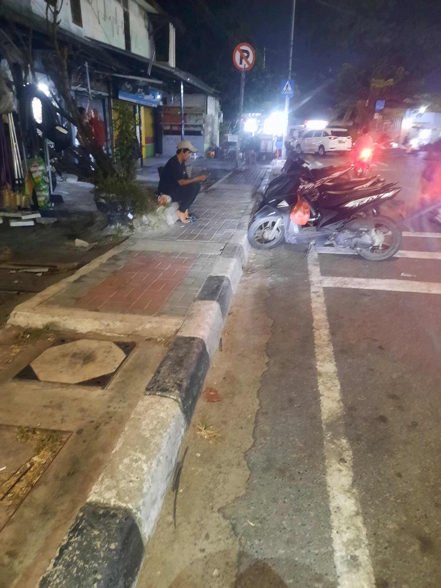 The image depicts a street scene at night in North Jakarta, Indonesia. A person wearing a dark shirt, dark pants, and a light-colored baseball cap is crouching on a sidewalk, looking at a mobile phone. In front of them is a red plastic bag. The sidewalk has decorative red tiles and a concrete curb painted with black and white stripes. Adjacent to the sidewalk is a road with a white lane marking. A black motorcycle is parked on the side of the road, with an orange plastic bag hanging from its handlebars. Other motorcycles and a white car are visible further down the street, along with streetlights and the facade of a building. A "no parking" sign is also visible. The overall lighting suggests it is nighttime.