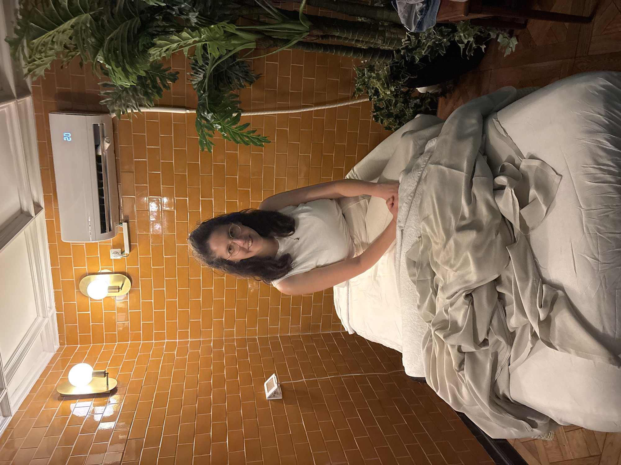 A young woman with dark hair and glasses, wearing a white t-shirt, is seated or leaning on a bed, smiling and looking directly at the camera. The bed is adorned with soft white and light grey bedding that appears rumpled. The room features distinctive walls covered in glossy, rectangular orange-brown subway tiles.
Above and to the left of the woman, a white air conditioner unit is mounted on the wall, displaying the number "20" in blue digital text. Below the AC, two modern wall sconces with circular diffusers and brass-colored oval bases emit a warm glow. A small, rectangular digital device is also affixed to the tiled wall further down.
Large, lush potted plants with broad green leaves are visible in the upper parts of the image and extending downwards, adding a natural and tropical aesthetic to the indoor space. Smaller potted plants with darker leaves are also present closer to the floor. A segment of a white-framed window or door is visible on the far left, and a wooden floor can be seen at the bottom. The warm artificial lighting from the sconces suggests an evening or nighttime setting. The woman appears relaxed and content.