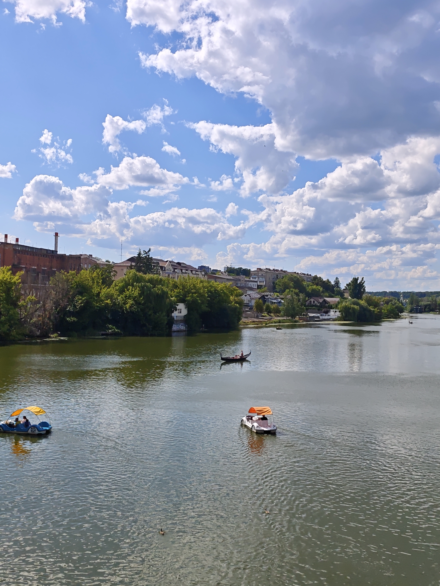 This is a landscape photograph taken on a sunny day with a partly cloudy sky, likely from a bridge or elevated viewpoint looking across a river. The river, appearing calm with ripples, occupies the majority of the frame. Several boats are visible on the water. In the foreground left, a small yellow paddleboat with an orange canopy carries at least two individuals. Further into the scene, slightly right of center, a gondola-style boat with a dark hull and a person in it is moving. To the right of the gondola, another paddleboat with an orange canopy is visible. A single duck is swimming in the water between the two foreground boats.
The background features a tree-lined riverbank with a mix of deciduous trees and some buildings nestled amongst them. On the left side of the riverbank, a large, old brick structure with a red-capped chimney dominates the skyline. Further along the bank, a cluster of residential buildings with various rooflines can be seen, suggesting an urban or suburban area. The overall impression is one of a recreational area within a city.
This scene is consistent with views along the Southern Bug River in the city of Vinnytsia, Ukraine, particularly areas known for recreational activities along the waterfront.