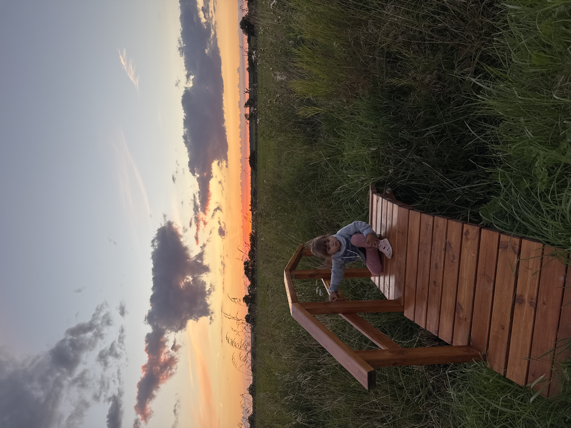 In Kempelevo, Russia, a young child sits on a wooden bridge that spans across a grassy field. The child is wearing a grey hoodie and pink patterned leggings. The setting appears to be outdoors during sunset, with the sky displaying hues of orange and pink, and scattered clouds. The bridge is constructed of polished wood and has a railing. The surrounding environment is a natural landscape with tall grasses and vegetation.