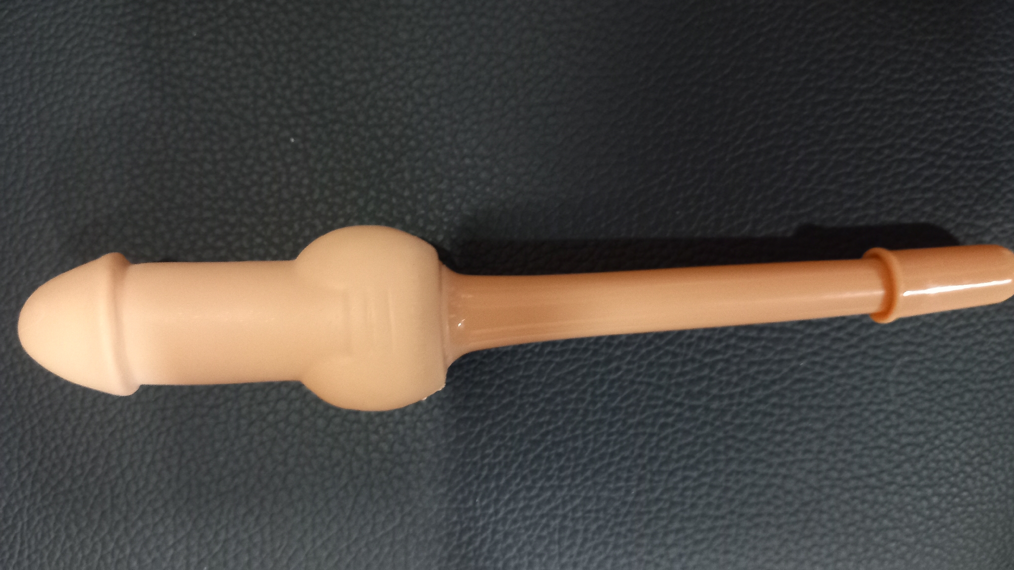 A close-up, high-angle, overhead shot shows a phallic-shaped, pale pink, plastic toy resting on a dark, textured surface, possibly faux leather. The toy is oriented vertically with its head pointing upwards. The toy features a rounded tip, a cylindrical shaft, a bulbous midsection with two raised bands, and a tapering lower shaft that ends in a flared, open base. The surface it rests on has a subtle, repeating pattern of small, raised dots and appears to be a dark blue or black color. There are no people or other objects visible in the frame. The lighting is even, casting soft shadows. The image appears to have been taken indoors. There is no indication of the location in the city of Kos, Greece.