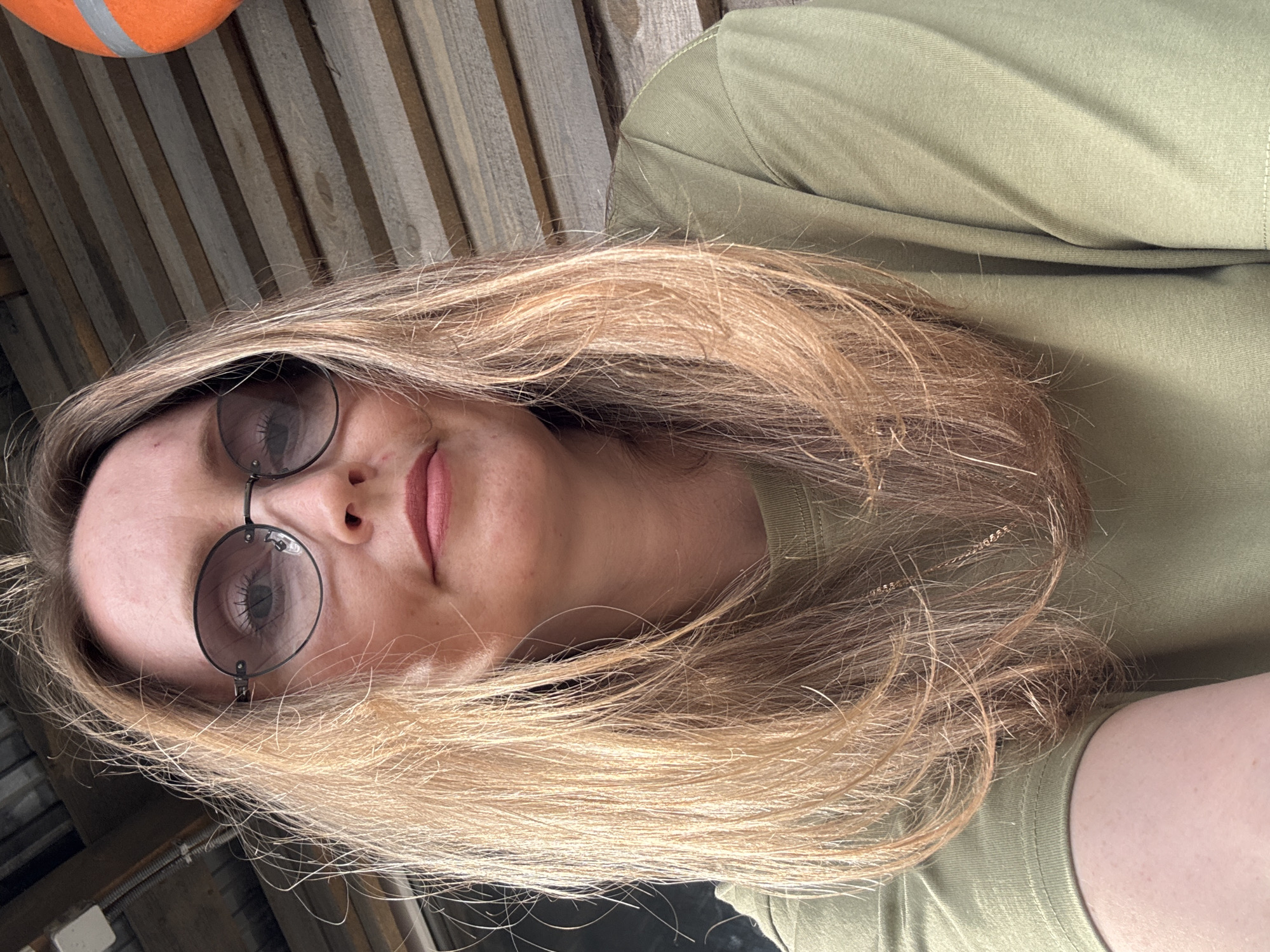 A close-up, chest-up photograph shows a woman with blonde hair and glasses. She is wearing a green t-shirt and is looking at the camera. The background appears to be a wooden structure, possibly part of a building or fence. There is an orange object with reflective stripes visible in the upper right portion of the image. The lighting suggests an outdoor setting.