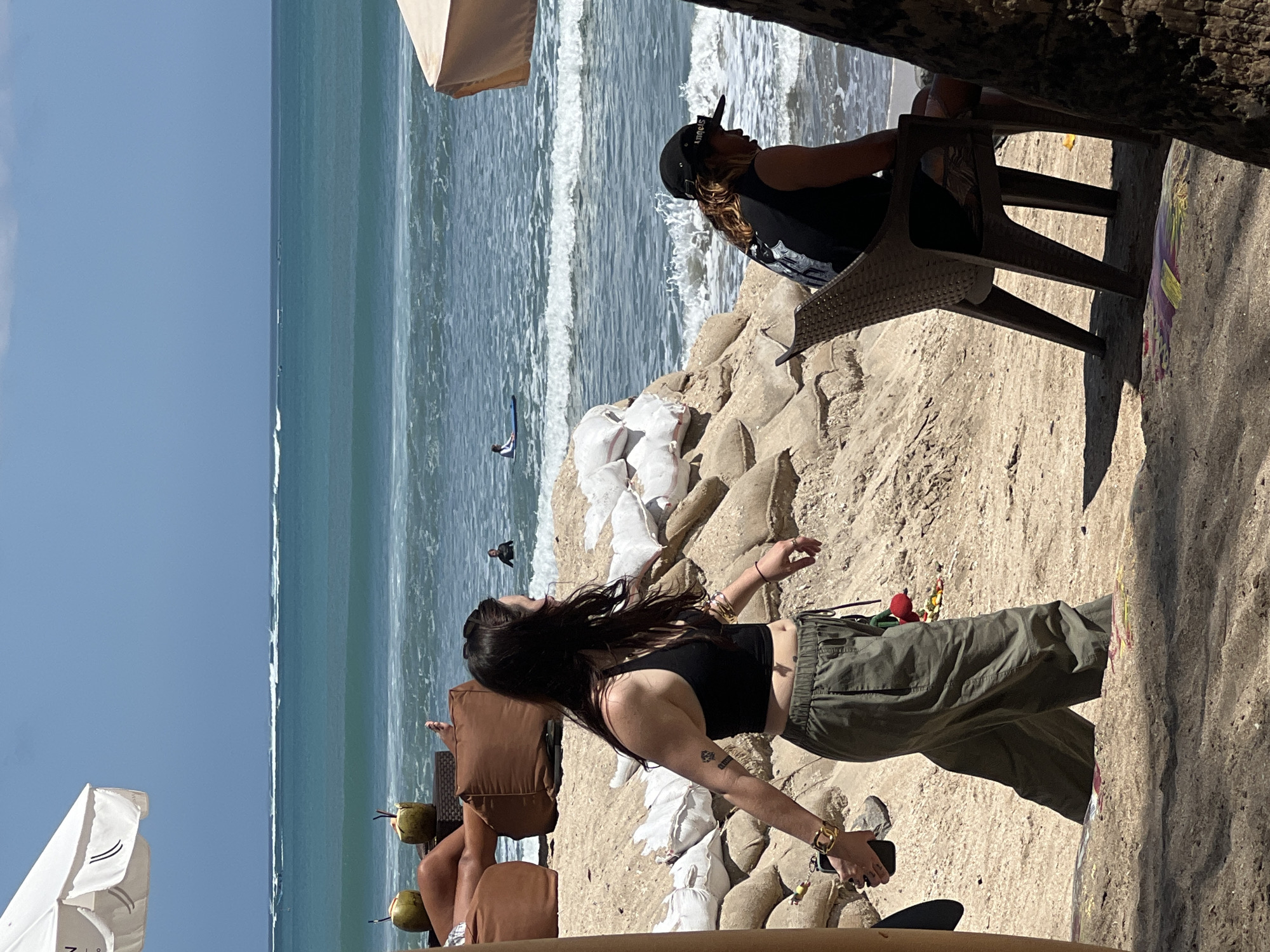 A medium shot captures a sunny day at Kuta Beach, Bali, Indonesia. The foreground shows a sandy beach with a woman in the middle ground walking from left to right. She wears a black tank top, olive green pants, and a gold bracelet on her left wrist. Her hair is long and dark, and she carries a dark smartphone in her left hand. Behind her, a man sits in a brown plastic chair facing right, wearing a black t-shirt and a black baseball cap. He is looking down. Behind the man, sandbags form a barrier against the ocean. The ocean is visible in the background with several surfers and a surfboard in the water. A white umbrella is visible in the top left corner. The overall scene depicts a typical beach day in Kuta, with people enjoying the sun and the ocean.