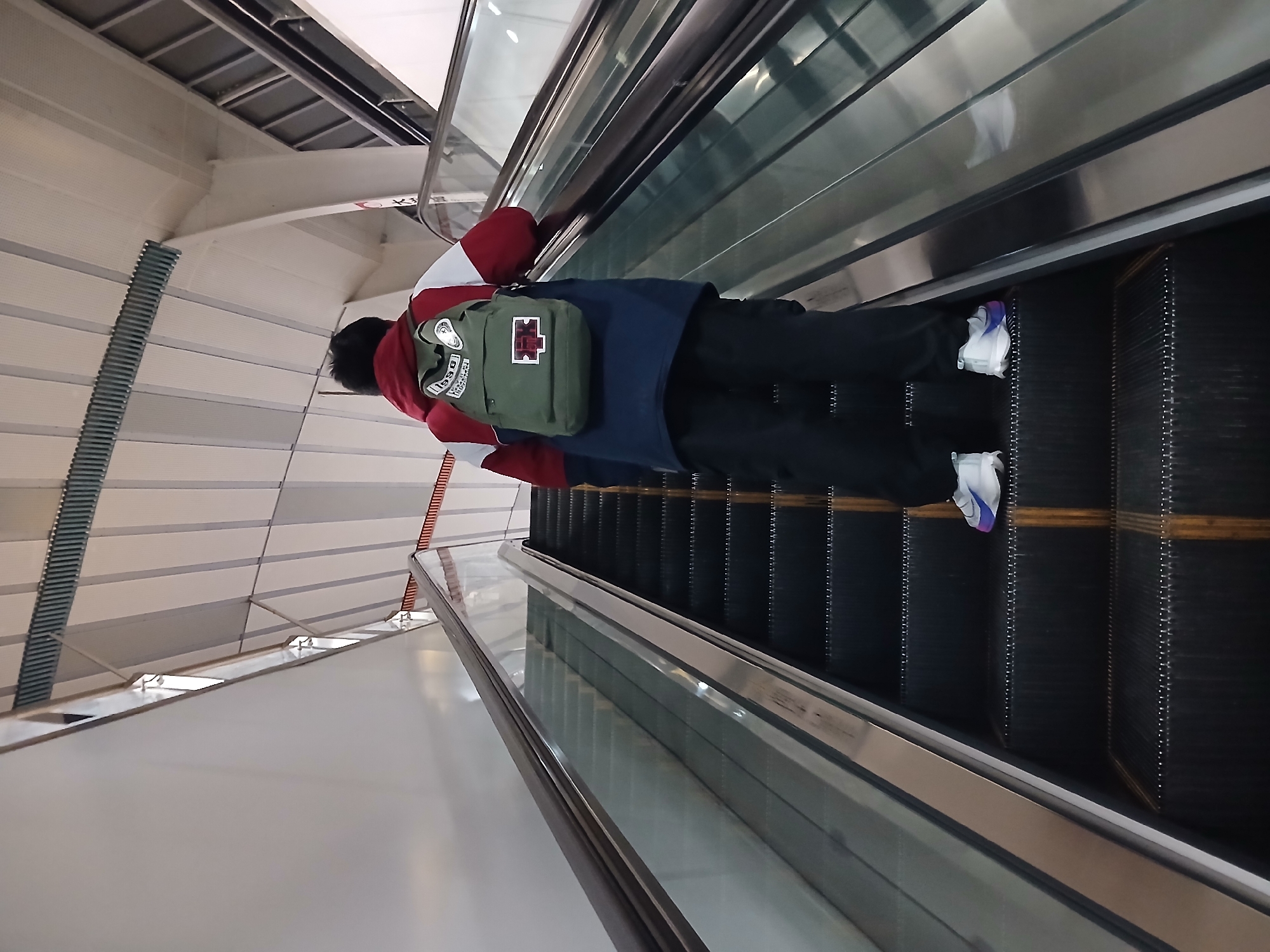 A person with a backpack is riding an escalator. The person is wearing a red and white jacket, dark pants, and light-colored sneakers with purple accents. The backpack is green with patches. The escalator has a stainless steel handrail and black steps. The location appears to be inside a modern building, possibly in the Chongqing High-tech Zone, China, based on visual architectural cues. The ceiling is white with linear design elements. The escalator is ascending.