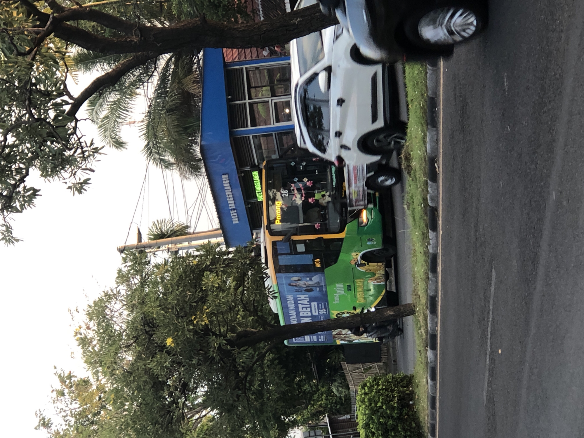 The image depicts a street scene in the city of Kludan, Indonesia, on a cloudy day. A bright green and yellow bus with advertisements on its side is the central focus. The bus has "trans jatim" written on it and a route number "28". In front of the bus, a small white trailer is attached. To the right of the bus, a white SUV is visible, and further to the right, the rear of a dark-colored car is seen. The background features a blue building with the words "HALTE TANGGULANGIN" and windows. Trees with green foliage and some yellow flowers frame the scene. Power lines are strung overhead. The road in the foreground is asphalt with white markings and a grey and black curb. A motorcycle is parked to the left of the bus. A grassy area with some scattered dry grass is located between the road and the bus.