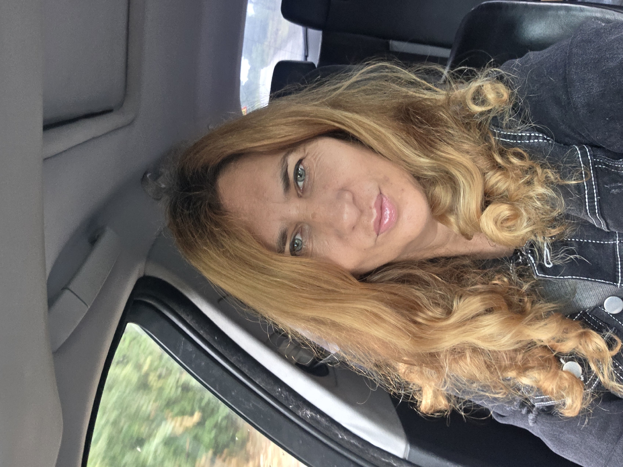 A medium-shot portrait captures a woman with long, wavy, light brown and blonde hair, and striking green eyes, smiling subtly at the camera. She appears to be taking a selfie or being photographed from a close distance, positioned inside a car. Her face is softly illuminated, suggesting daylight.
She is wearing a dark garment, possibly a denim jacket or shirt, detailed with prominent white stitching and silver buttons on the front. Her lips are a light pink, possibly accentuated with lip gloss.
The setting is the interior of a vehicle. Elements of the car visible include a grey sun visor above, a dark headrest, and part of a car door frame and window to her left. Through the car window, a blurred background of green foliage and hints of light indicate an outdoor environment, possibly a road or natural landscape. The overall mood of the portrait is calm and pleasant.
The location context for this image is Kamenar, Bulgaria. No text is visible in the image.