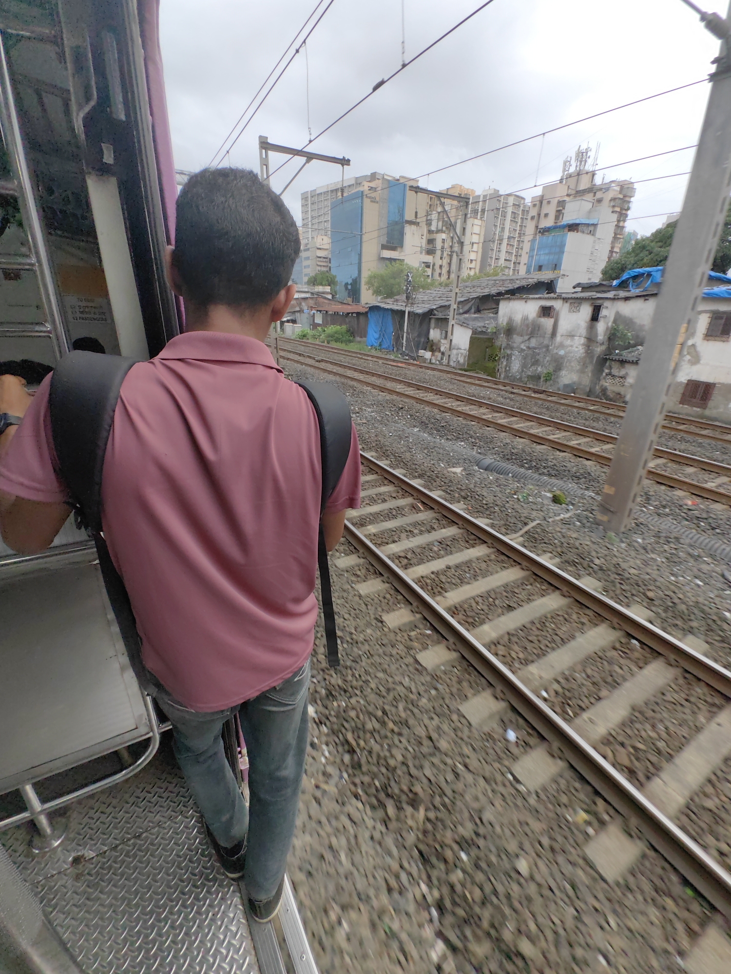 The image captures a person, from the back, standing near the open door of a train carriage, with a backpack slung over their shoulders. The person is wearing a pink polo shirt and blue jeans. The train is adjacent to a set of railway tracks that run alongside a densely populated urban area in Mumbai, India. The background reveals a mix of residential buildings, some appearing to be high-rise apartments, alongside smaller, older structures and makeshift dwellings. The sky is overcast. The scene suggests movement and the typical environment of a commuter train route in Mumbai, likely a suburban railway line.