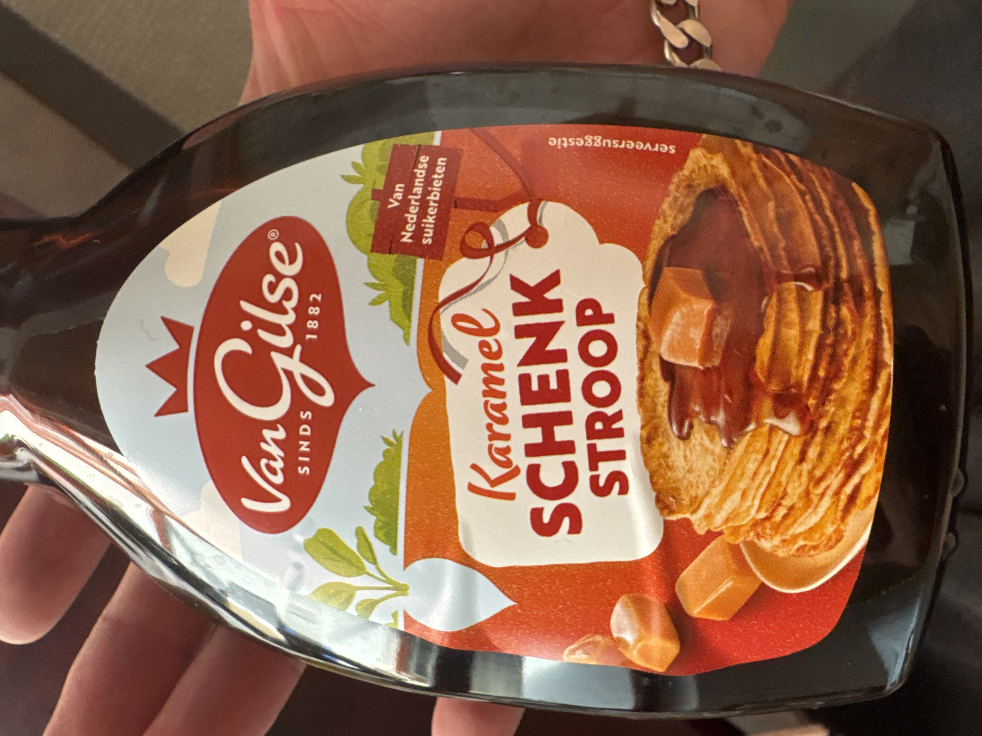 A dark brown bottle of Van GIlse Karamel Schenk Stroop is held by a person's hand. The bottle has a white and red label with a crown logo. Text on the label reads "Van GIlse SINDS 1882" and "Karamel SCHENK STROOP". Below the text, there is an illustration of pancakes drizzled with syrup and caramel pieces. A small red sign on the label says "Van Nederlandse suikerbieten". The scene is not in a city.
