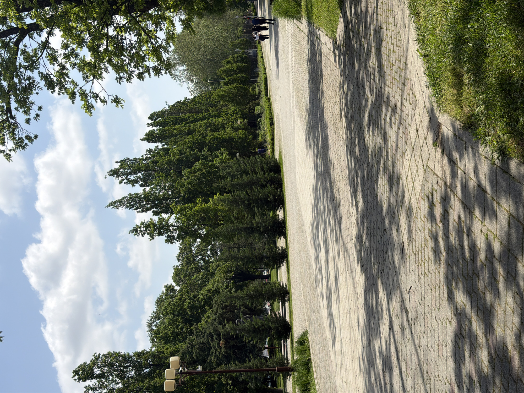 The image depicts a brick-paved pathway lined with trees on either side in a park setting. The location is Krasnodar, Russia. Sunlight filters through the trees, creating shadows on the path. A group of people are walking in the distance. The sky is visible through the trees, with some clouds present. A street light fixture is also visible.
