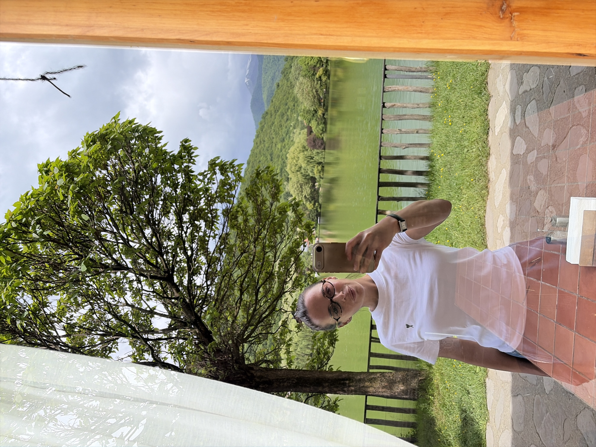 A woman is taking a selfie in a location with a view of a lake or large body of water. The setting appears to be outdoors, possibly from a building or structure that offers a framed view of the natural landscape. A large tree with green leaves is visible to the left of the woman. In the background, there is a body of water, a wooden fence running along the water's edge, a green grass area, and a forested hillside. In the distance, there is a mountain range. The woman is holding a smartphone up to take the photo and is wearing glasses and a white t-shirt. The setting could be in or around Napareuli, Georgia, considering the landscape aligns with the region's characteristics.