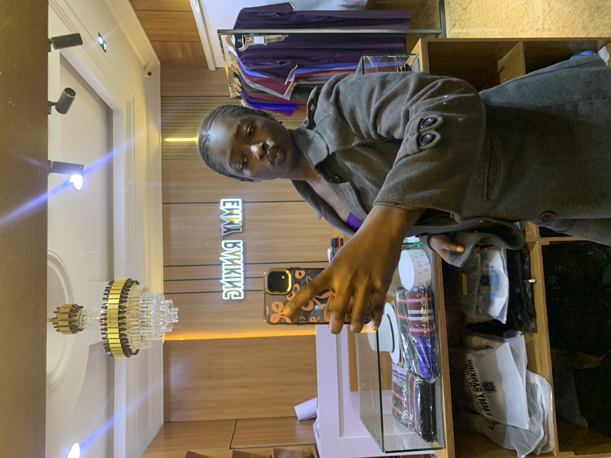 In an indoor retail space, likely a boutique or clothing store, a young Black woman is taking a selfie in a mirror. She is wearing a dark grey coat over a purple top. The woman is holding a smartphone in her right hand, angled to capture her reflection. The background reveals a well-lit environment with wooden paneling on the walls and shelves displaying clothing and accessories. A prominent chandelier hangs from the ceiling, and neon signage in reverse, reading "EMMY RANKING," is visible on the wall behind her. To the right, clothing items are neatly displayed on a rack. The overall impression is of a fashionable establishment.