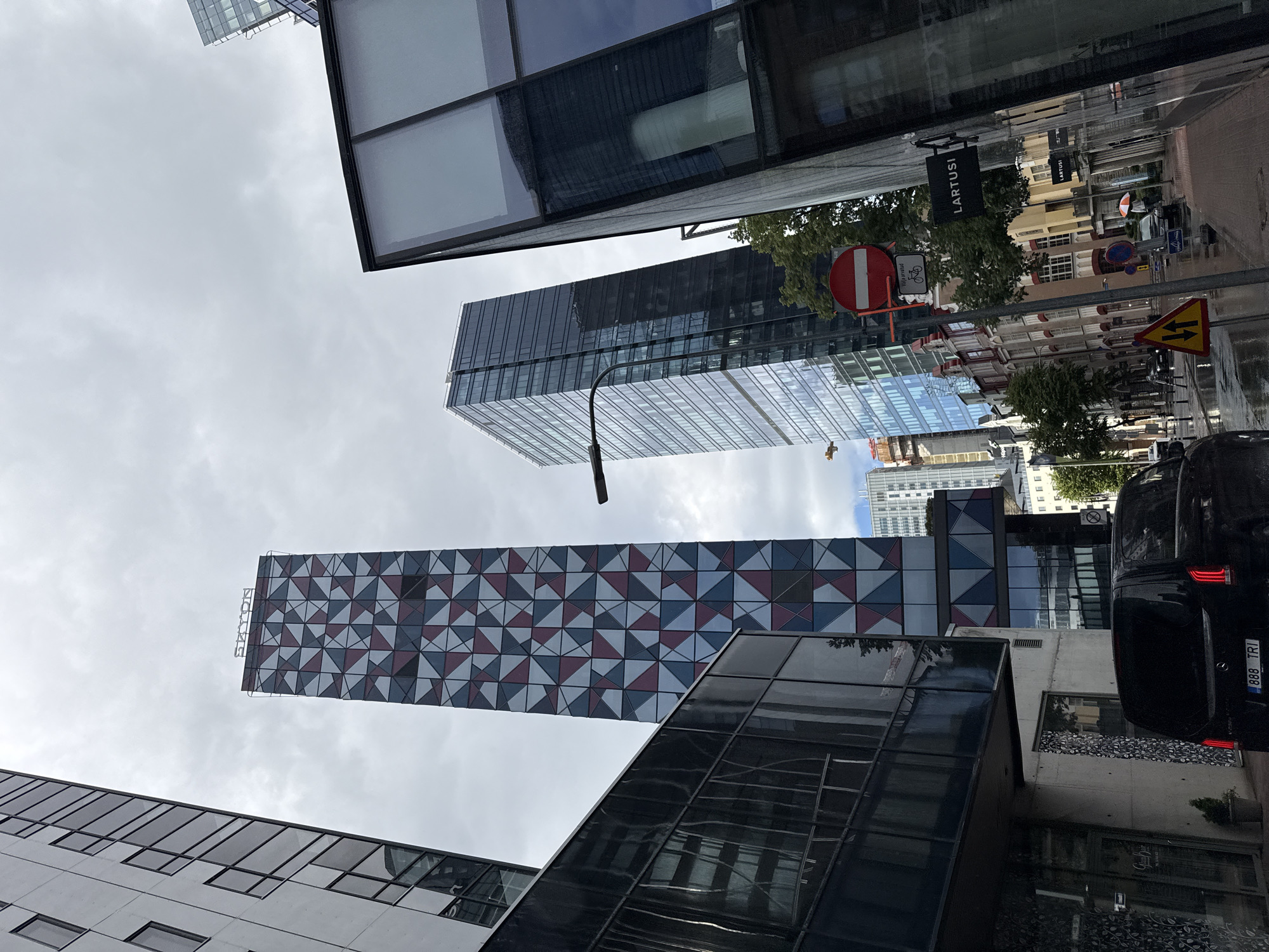 In this eye-level, wide-angle shot, the scene captures a street view in Tallinn, Estonia, under an overcast sky. The foreground is dominated by the angular, dark glass facade of a modern building on the right, reflecting the surrounding environment and sky. To the left, a black minivan with a license plate reading "888 TRI" is parked.
Further into the scene, several contemporary high-rise buildings stand tall. One prominent skyscraper features a striking geometric facade composed of red, blue, and gray triangles, topped with the word "SEAPORT" in white lettering. Adjacent to it is another sleek, glass-covered skyscraper. In the middle ground, a row of older, colorful buildings with arched windows lines the street, contrasting with the modern architecture.
Traffic signs are visible, including a circular red and white "no entry" sign with a bicycle symbol below it, and a yellow triangular warning sign with two black arrows pointing left and right. A street sign for "LARTUSI" is mounted on a building. The street surface appears wet, suggesting recent rain. A few pedestrians are present, one holding a red and white umbrella. Trees with green foliage add natural elements to the urban landscape. The overall atmosphere is one of a modern European city with a mix of historic and contemporary structures.