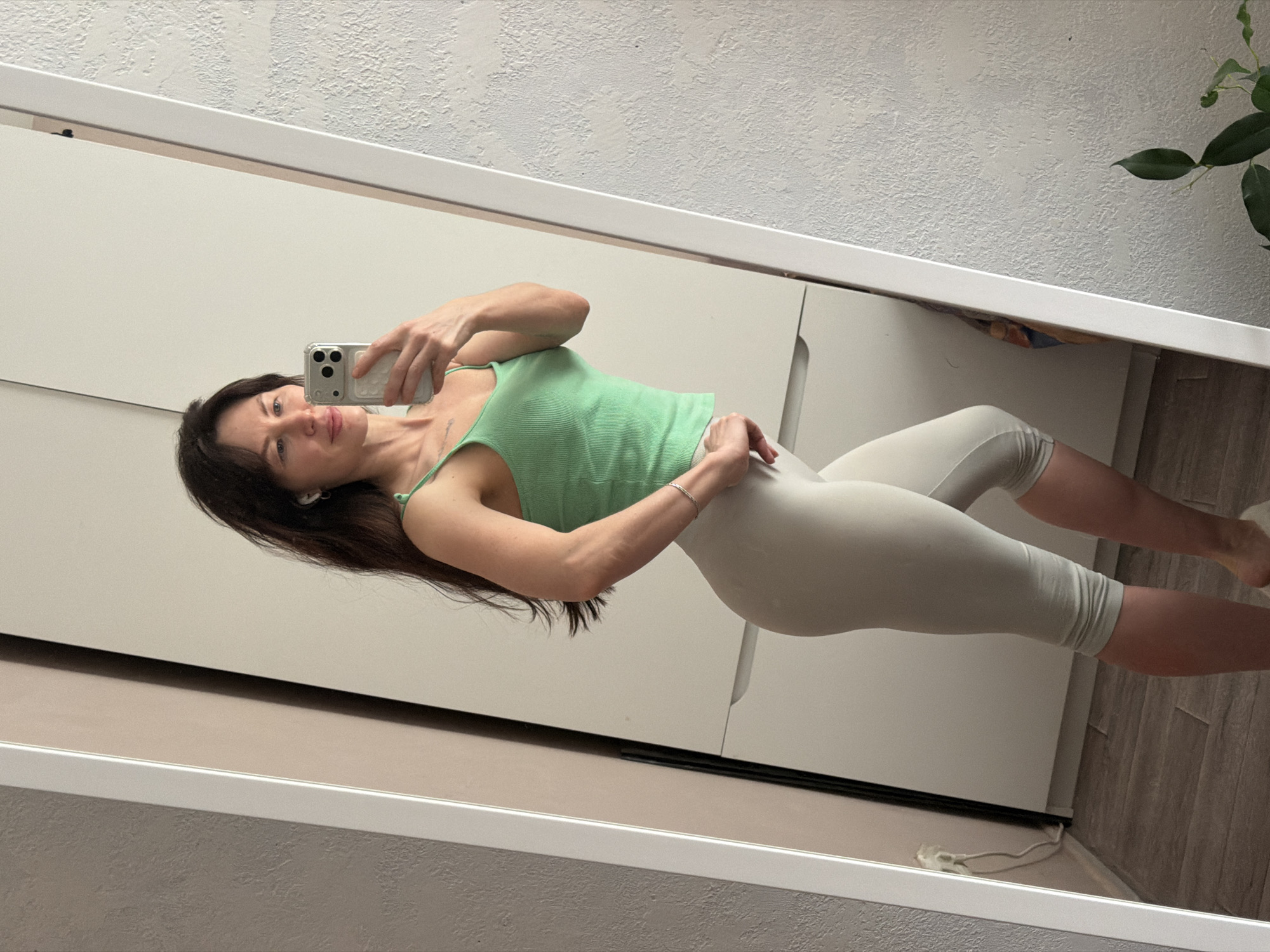 A full-body selfie of a woman in workout attire is reflected in a mirror. She is wearing a light green tank top and light-colored workout leggings. Her dark hair falls over her shoulder. She is holding a smartphone, using it to take the picture. The background shows a white wardrobe, a textured wall, and a partial view of a plant. The location is likely indoors. The city of Perm, Russia is not discernible from the image.