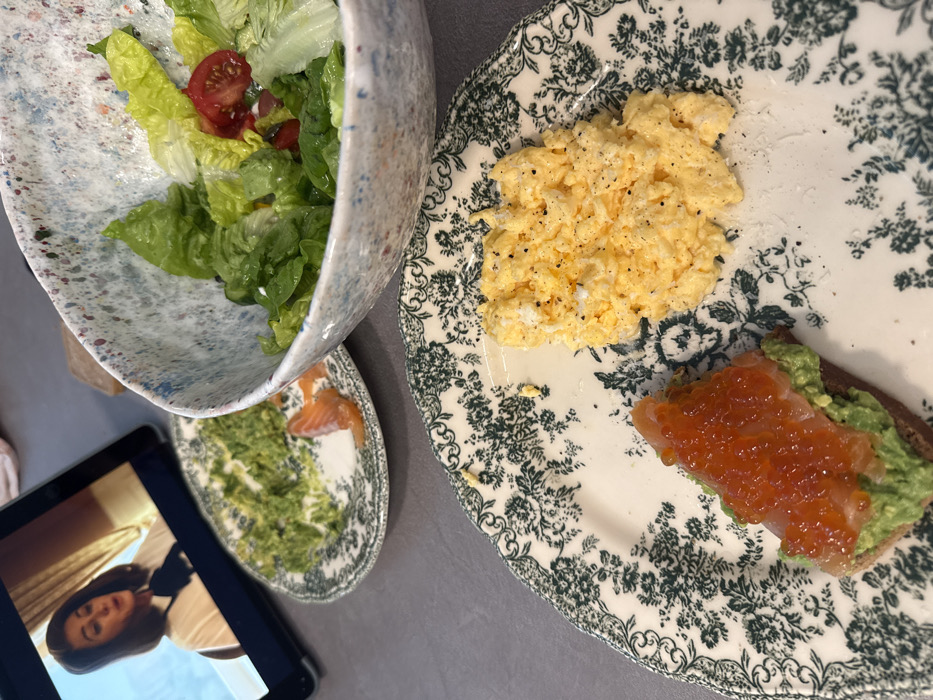 The image displays an indoor setting, likely a private residence, with a meal served on a gray countertop. In the foreground, a white ceramic plate with a dark green floral pattern holds a portion of yellow scrambled eggs seasoned with black pepper, and a piece of dark toast topped with mashed green avocado and bright orange fish roe. Adjacent to it on the right, a large, speckled ceramic bowl contains a green salad consisting of lettuce leaves and sliced red cherry tomatoes. Further back on the left, a smaller, similarly patterned plate shows remnants of mashed green avocado and several thin slices of pink smoked salmon. In the upper left background, a black electronic tablet is playing a video, depicting a woman with dark hair wearing a white shirt with a black bow, against a background of light-colored curtains. The visual information available does not provide specific details to identify a precise location within Tel-Aviv, Israel.