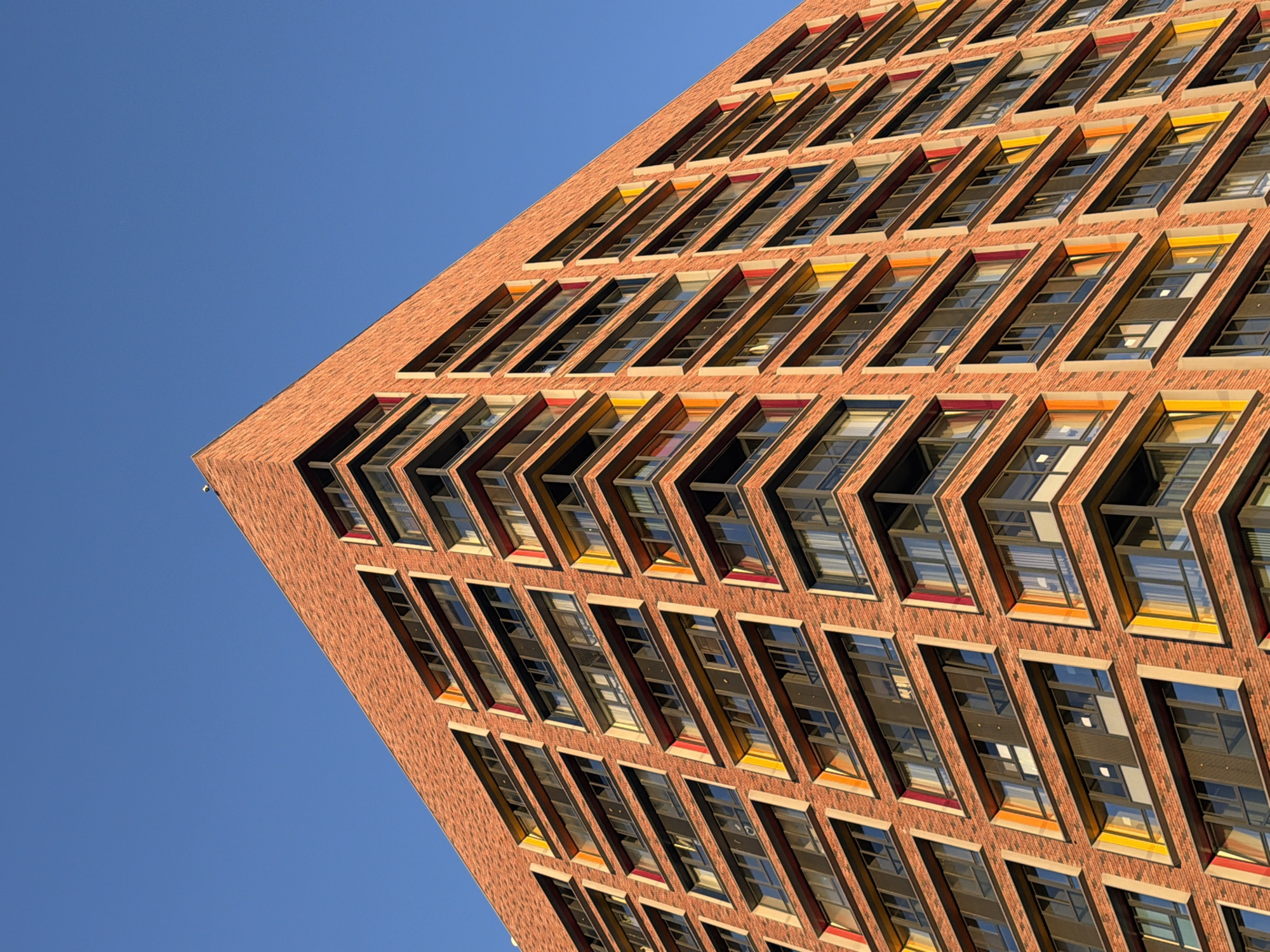 An upward, exterior, eye-level, full shot captures the exterior of a modern, multi-story apartment building constructed from reddish-brown brick. The building has a distinctive architectural design featuring alternating colored window frames in yellow and reddish-orange. The windows are dark-paned, and some have visible balconies. The building is set against a clear, bright blue sky, indicating daytime. The scene is devoid of any people or vehicles. The low angle emphasizes the scale and height of the building.