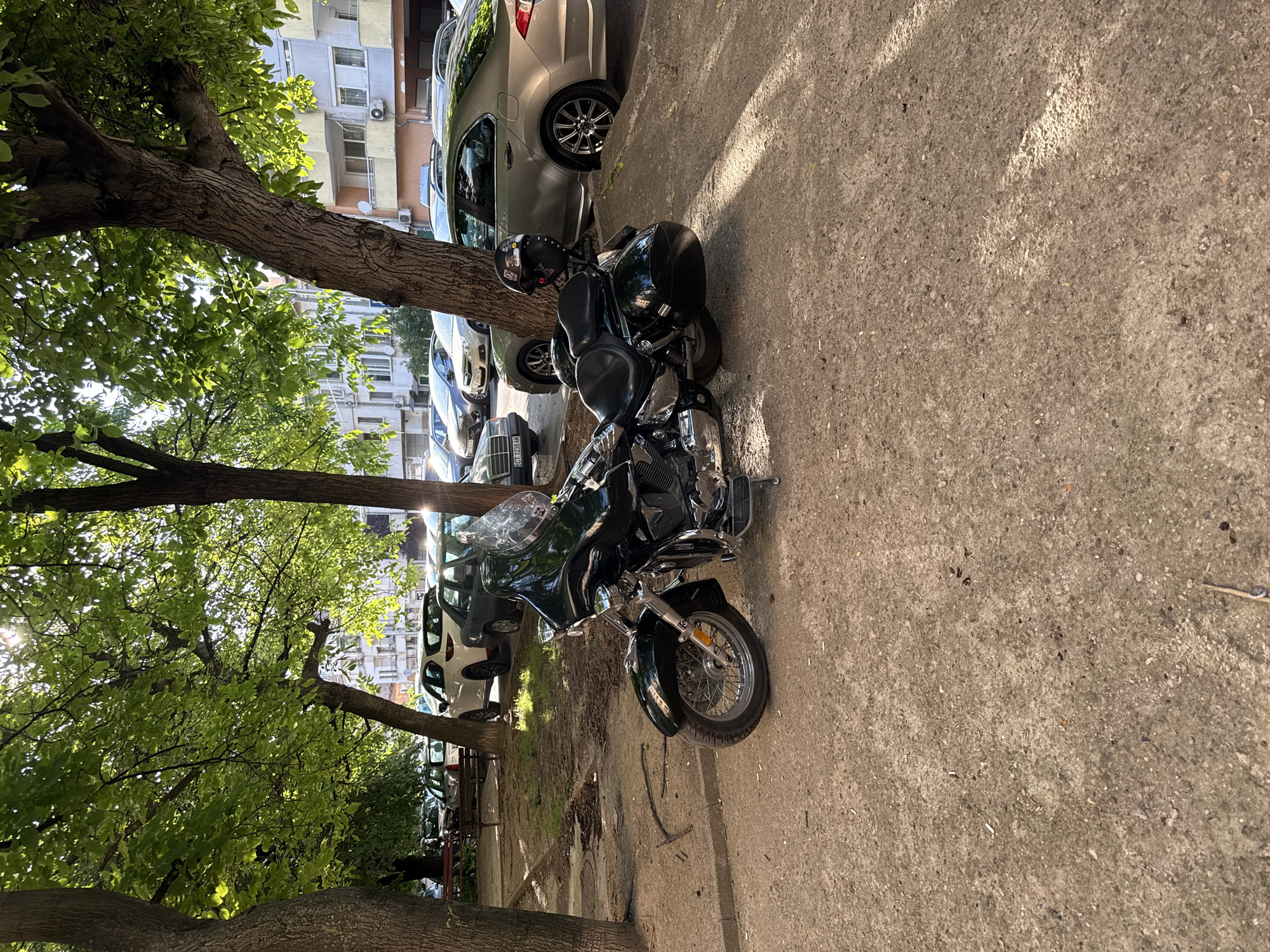 The image depicts a street scene in Stara Zagora, Bulgaria, on a sunny day. In the foreground, a dark green motorcycle is parked on a concrete surface. The motorcycle is a cruiser style, featuring a large windshield, saddlebags, and chrome detailing. Behind the motorcycle, several cars are parked along a curb. These include a light silver sedan, a dark sedan, and a white SUV. The background shows a multi-story apartment building with balconies. Trees with lush green foliage line the street, casting dappled shadows on the ground. The overall impression is a typical urban street with parked vehicles and residential buildings.