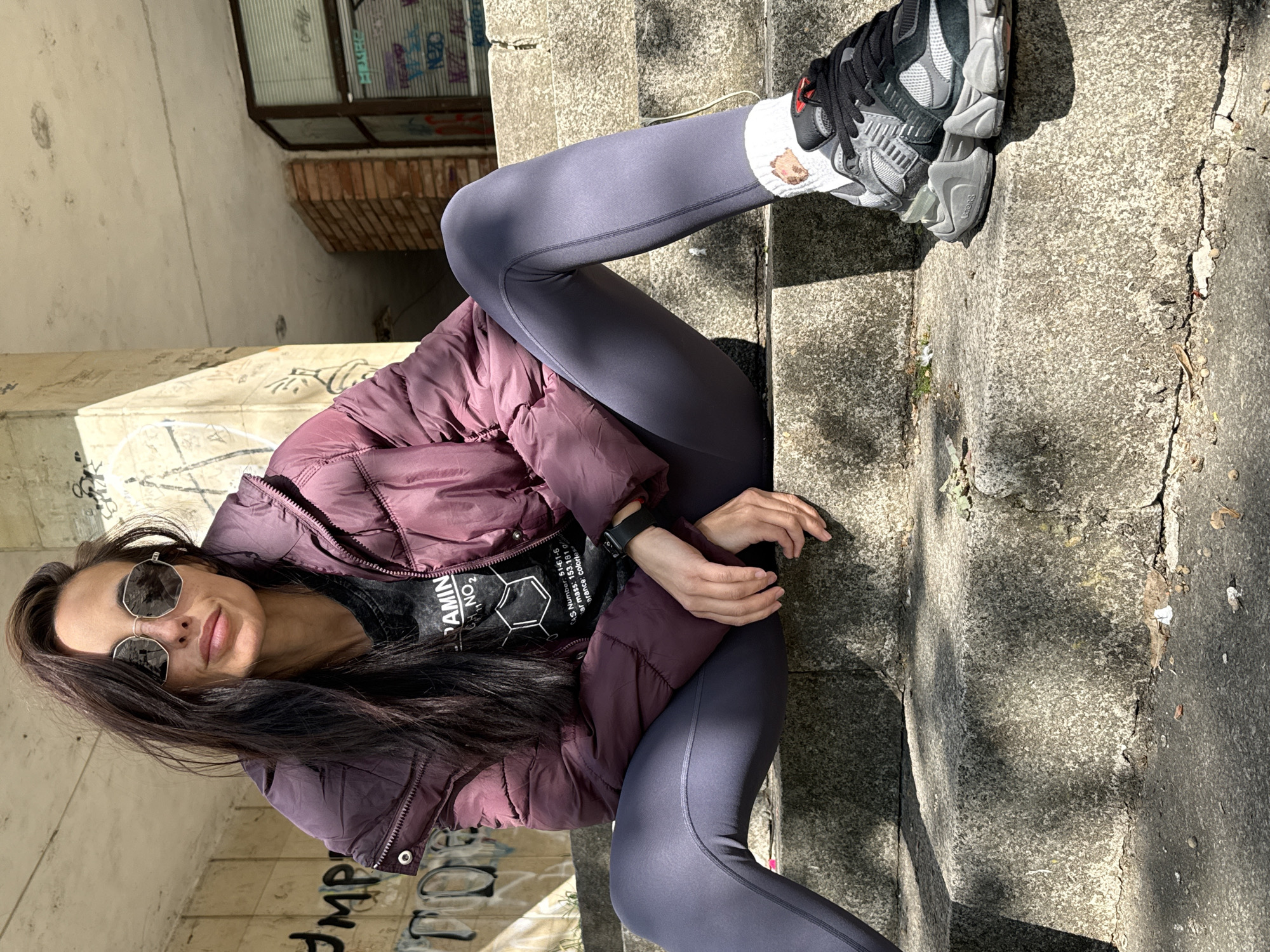 The image shows a woman sitting on concrete stairs. She is wearing a maroon puffer jacket over a black t-shirt, grey leggings, and grey and black athletic shoes with white socks. She has dark hair and is wearing sunglasses. The stairs are part of a concrete structure, possibly a building or underpass, and there is graffiti on the walls. The location is Pleven, Bulgaria.
