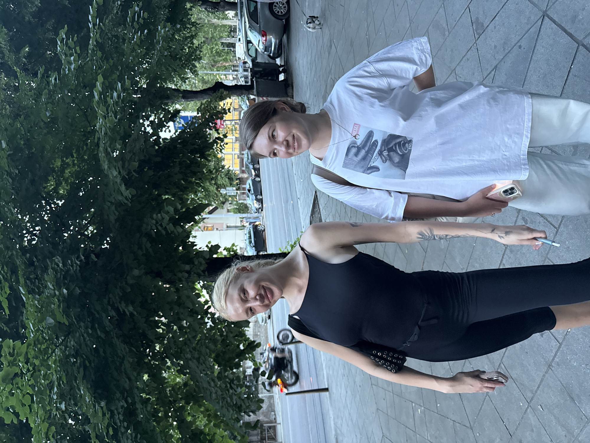 Two women stand on a tiled sidewalk next to a street. One woman wears a white t-shirt with a graphic and white pants. The other woman wears a black tank top and black pants. Trees are visible on the left side of the image. A street with vehicles and buildings is behind them. A motorcycle is visible on the street. The location is likely in Belgrade, Serbia.