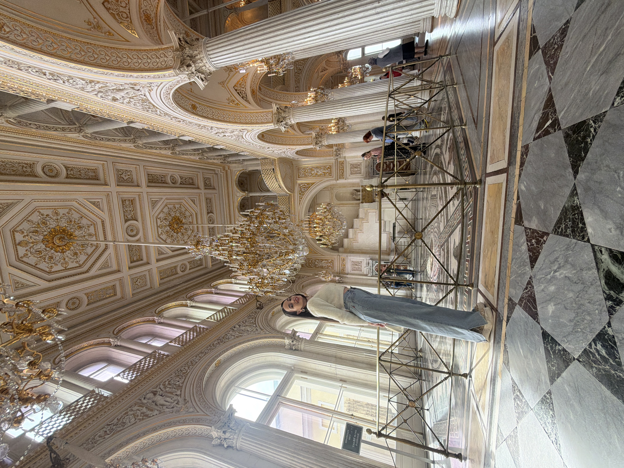 Here is a description of the image:
A woman in a white sweater and jeans stands in an ornate hall. She is facing the camera with her body slightly turned to the left. The hall is decorated in gold and white, with chandeliers, columns, and detailed molding on the ceilings and walls. A metal barrier is in front of her, and other people are seen behind the barrier. The floor is a checkerboard pattern of black and gray marble tiles. Large windows are visible in the background, letting in daylight.
Based on the architectural style and the prevalence of gold decoration, it is likely that this photo was taken at the Winter Palace in St. Petersburg, Russia.