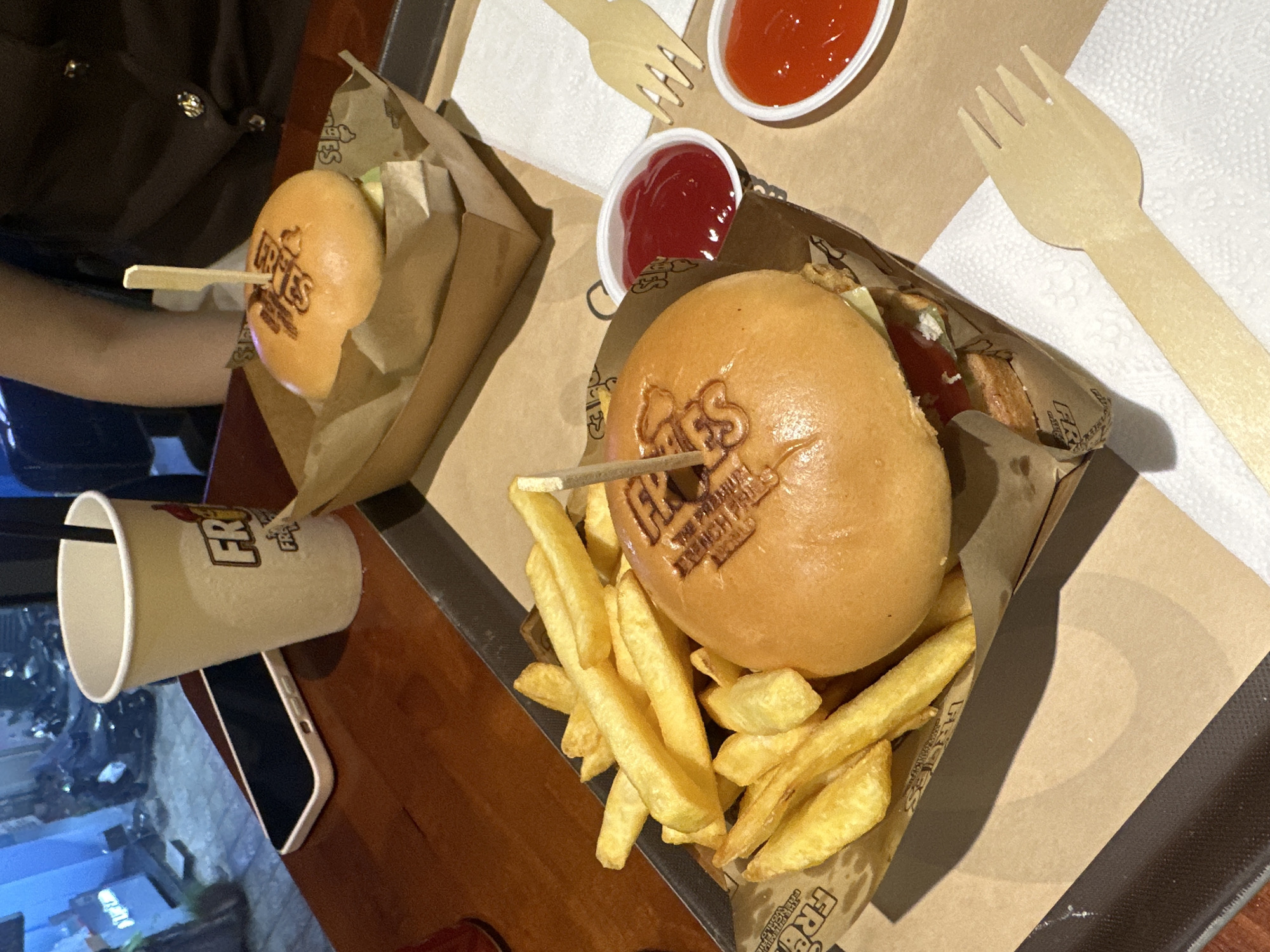 The image shows a close-up, top-down view of a fast-food meal. Two burgers, each secured with a wooden skewer and branded with "Frries" text, are presented in brown paper containers. The burger on the right is accompanied by a generous serving of french fries. Each burger container has a small plastic cup of red sauce (likely ketchup) nearby. Wooden forks rest on white napkins. A white paper cup with "Frries" branding is visible in the lower-left corner, along with a white-cased mobile phone. An individual wearing a dark-colored garment is partially visible in the upper-left. The setting appears to be a table, with the background hinting at an urban environment visible through a window, possibly in Thủ Đức, Vietnam. The meal is presented on a dark tray.