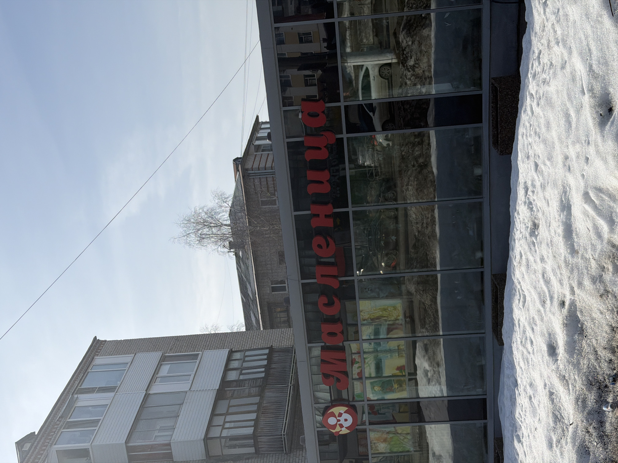The image depicts a street scene in what appears to be Berezniki, Russia, based on the presence of the Cyrillic word "Масленица" (Maslenitsa) on a building facade. The view is taken from a low angle. The "Maslenitsa" lettering is large and red. A red logo is beneath the word "Maslenitsa." A multi-story building is to the left of the "Maslenitsa" building. The "Maslenitsa" building has large windows. The windows reflect other buildings and cars. Patches of snow cover the ground to the right. The sky is partially cloudy. A wire stretches diagonally across the sky.