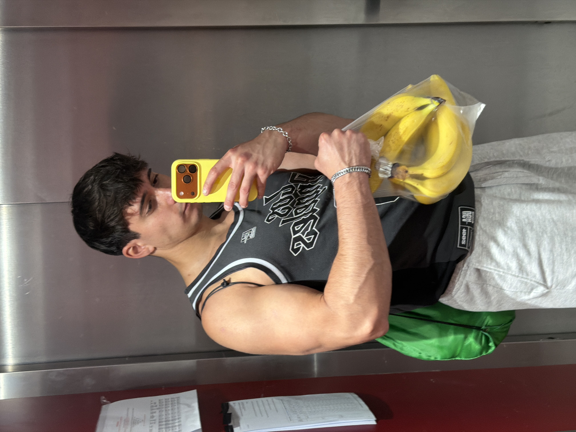 A young man is captured in a mirror selfie, holding a bunch of ripe yellow bananas in a clear plastic bag in his left hand and a yellow smartphone (likely an iPhone) in his right. The image is rotated 90 degrees clockwise, showing him standing sideways in an indoor setting in Sofia, Bulgaria.
The man has dark, short, wavy hair and an athletic physique, with noticeable bicep and forearm musculature. He is wearing a black sleeveless athletic tank top with white trim and white stylized graphics on the front, including the number "23" and other partial text, reminiscent of a basketball jersey. Light grey sweatpants are visible below his top. He accessorizes with two silver-colored bracelets, one a chain-link on his left wrist and another round-link style on his right. A bright green fanny pack or small bag is tucked at his hip.
The background consists of vertical brushed metallic panels, suggesting an elevator or an institutional hallway. In the lower foreground, a dark red ledge or counter holds two stacks of papers or documents, one of which is clipped to a board; text on these documents is illegible due to distance and angle. The man is focused on his phone screen as he takes the photo. The bananas are vibrant yellow, indicating ripeness. No specific time of day or weather cues are evident beyond the indoor lighting.