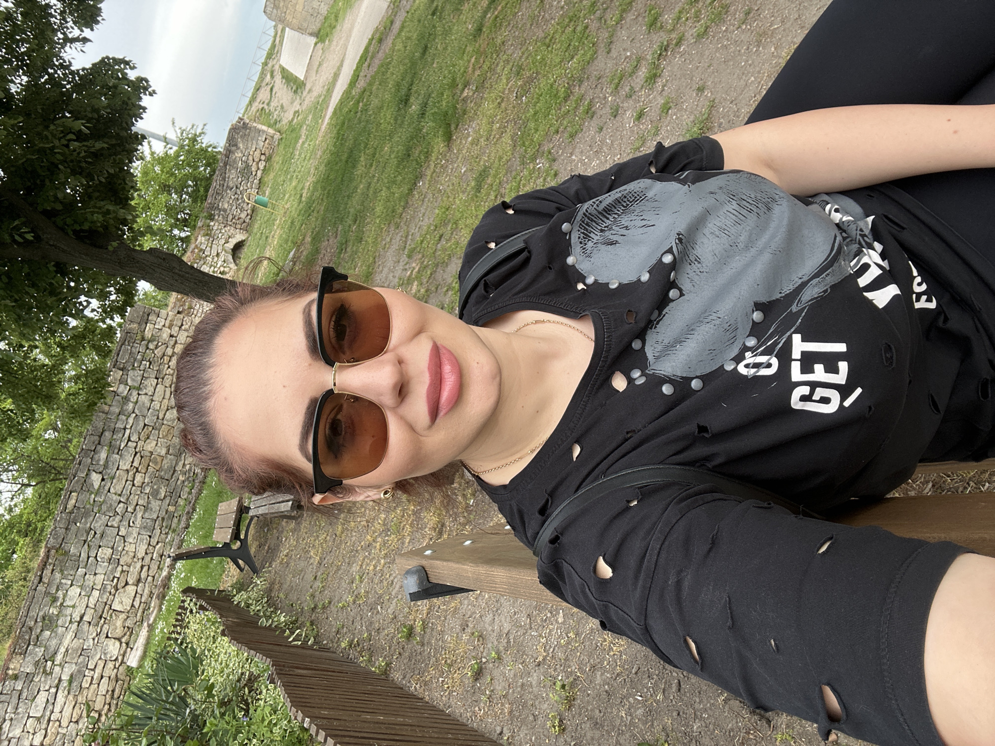 A selfie shows a woman lying on a wooden bench outdoors. She has auburn hair pulled back in a ponytail, brown sunglasses, and pink lipstick. She wears a black t-shirt with holes and an image of a face with studded details. The word "GET" is visible on the t-shirt. She also wears black leggings. The background shows a grassy area, a stone wall, trees, and a wooden fence. The image was taken in Vidin, Bulgaria.