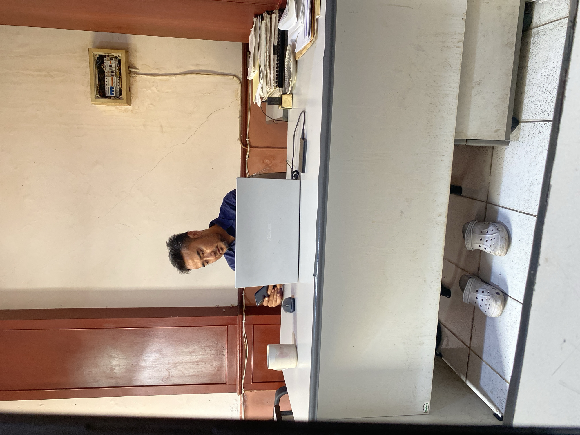 A man is sitting at a desk in what appears to be an office in North Jakarta, Indonesia. He is looking at his smartphone, which he is holding in his left hand. A laptop is open in front of him on the desk, and a computer mouse is to the left of the laptop. A white mug with a handle is next to the mouse. A stack of papers is on the right side of the desk. There are two pairs of white Crocs shoes under the desk. The walls are a light color, and there is a dark wooden cabinet to the left. An electrical panel is mounted on the wall to the right. The overall impression is one of a simple, functional workspace.