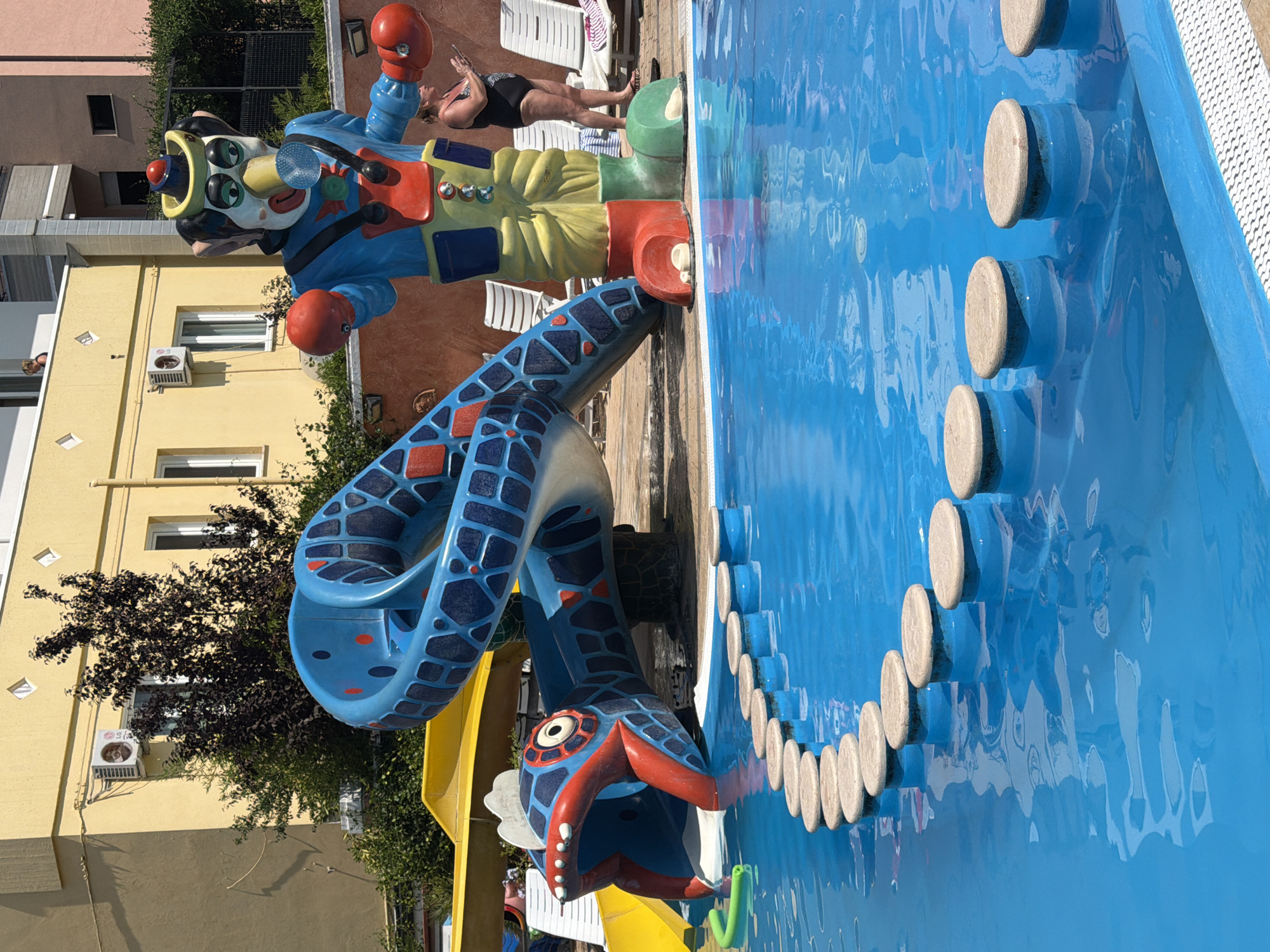 In Gatteo, Italy, this image captures a vibrant outdoor swimming pool area at a water park or resort. Dominating the scene is a large, colorful water slide designed to resemble a snake with a gaping fish-like mouth serving as the entrance. Beside the slide stands a giant clown statue, its red boxing gloves extended. A woman in a black swimsuit is visible in the background, looking at her phone. The pool itself is a brilliant blue, with a line of circular stepping stones creating a path across the water's surface. In the background, a light yellow building with visible air conditioning units and windows suggests the presence of accommodation or facilities. Green bushes and trees surround the area, and lounge chairs are placed on the tiled perimeter of the pool. The overall atmosphere is one of summer fun and recreation.