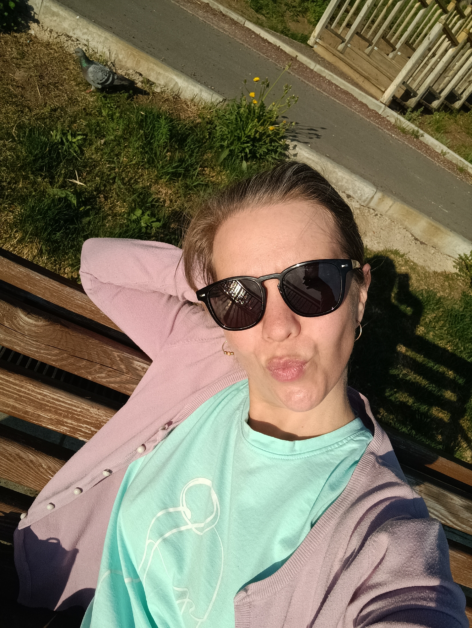 A woman is sitting on a wooden bench, seemingly taking a selfie. She is wearing sunglasses, a light pink cardigan, and a light blue t-shirt with a graphic design. Her arms are behind her head, and she is puckering her lips. A pigeon is visible in the upper left corner of the frame. The bench is next to a grassy area and a paved walkway, suggesting a park setting. Based on the details, it's likely the city of Chaikovsky, Russia, although specific landmarks are not visible to confirm this definitively.