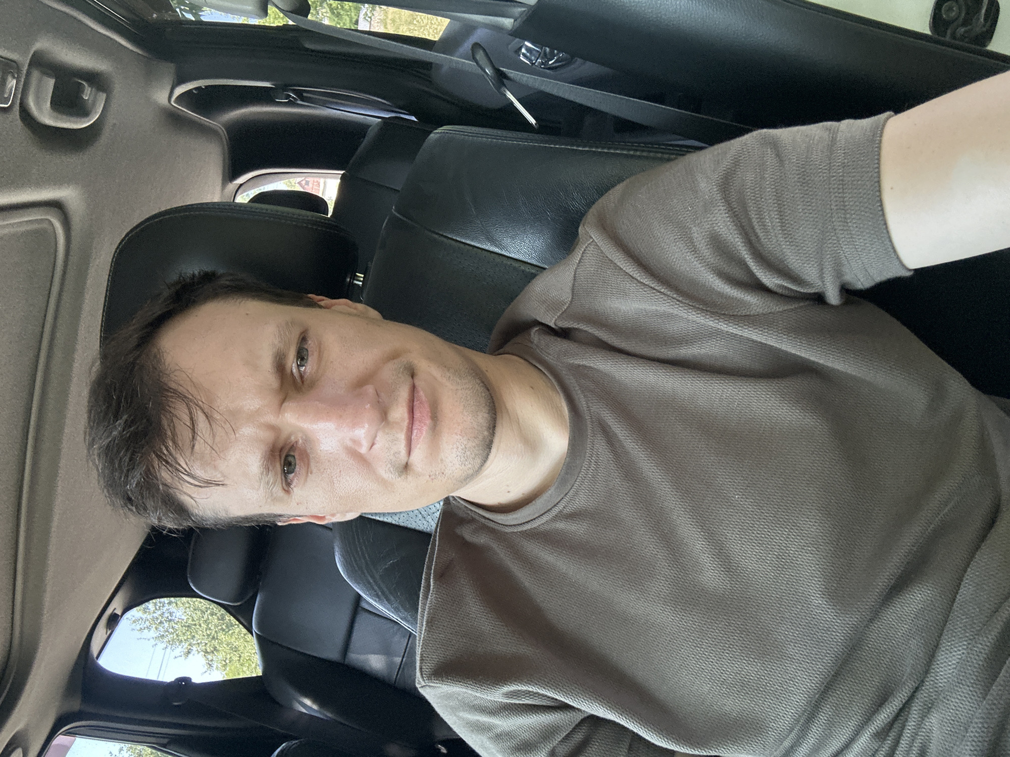 A man with short, dark hair and a neutral expression is taking a selfie from the driver's seat of a vehicle. He is wearing a brown, textured t-shirt. The interior of the car is visible, with black leather seats and a sunroof. Outside the windows, trees and foliage can be seen, suggesting a location with greenery. The image appears to be taken during the daytime.