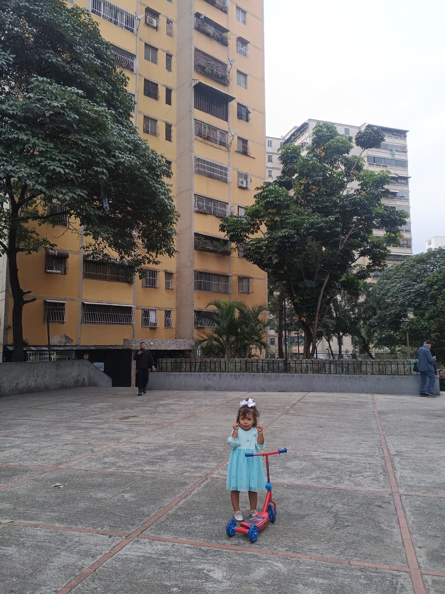 The image captures an outdoor scene within a residential area, likely a plaza or courtyard, set against a backdrop of multi-story buildings and lush greenery in Caracas, Venezuela.
The primary subject is a small child, likely a girl, positioned in the lower-left center of the frame. She wears a light blue dress with white patterns, white shoes, and a white bow in her dark, curly hair. She stands next to a red and blue three-wheeled kick scooter and is making a gesture with both hands raised, fingers slightly curled.
In the background, two adult males are visible. One man, dressed in dark clothing, is walking on the left side of the frame, approaching the entrance of a building. Another man, wearing a blue shirt and jeans, stands near a metal fence on the right side.
The setting is characterized by several tall, residential apartment buildings, predominantly light yellow or cream-colored. These buildings feature numerous windows, many of which are fitted with security bars or grates, and some have small balconies. Air conditioning units are visible on several facades. Large, green leafy trees are scattered throughout the scene, some partially obscuring the buildings, adding a natural contrast to the urban structures. Palm trees are also visible behind a fence on the right.
The ground is a paved concrete surface, divided into a grid pattern by reddish-brown lines. The sky appears overcast, suggesting a cloudy day with diffused light, but it is clearly daytime. No discernible text is visible in the image. The overall impression is a candid moment from everyday life in an urban residential environment.
