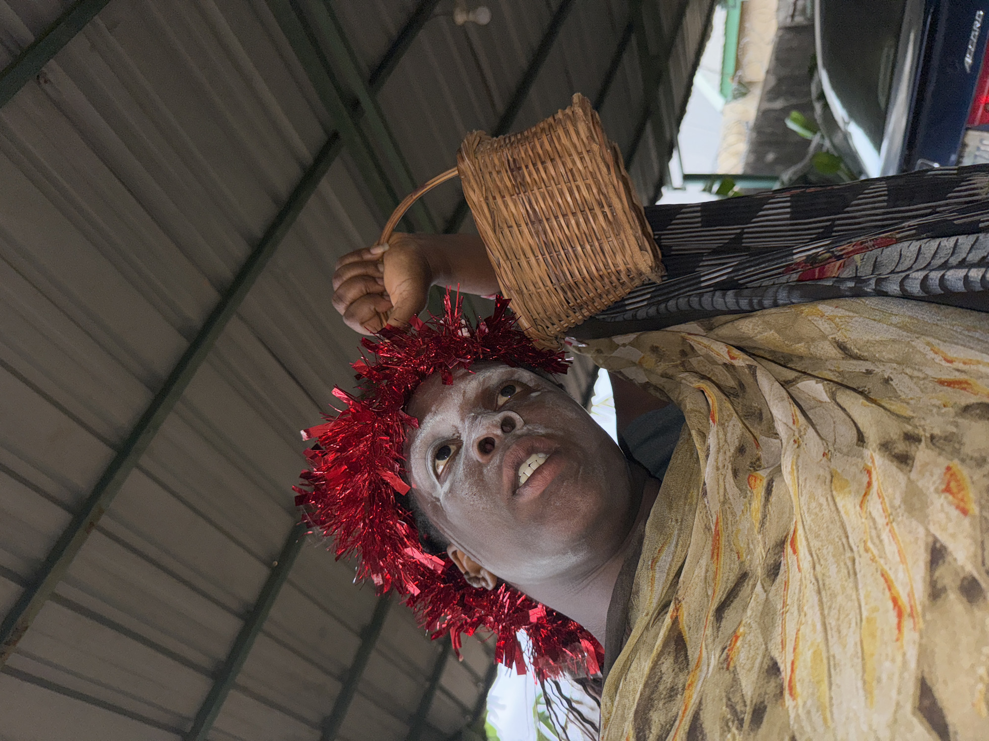A low-angle shot shows a person of dark complexion, with face painted white, wearing a red tinsel garland around their head and draped in a light-colored patterned shawl. They are holding a small woven basket by its handle above their head. The background includes a corrugated metal roof, parts of a vehicle, and foliage. The location is likely Sabon Tasha, Nigeria.
