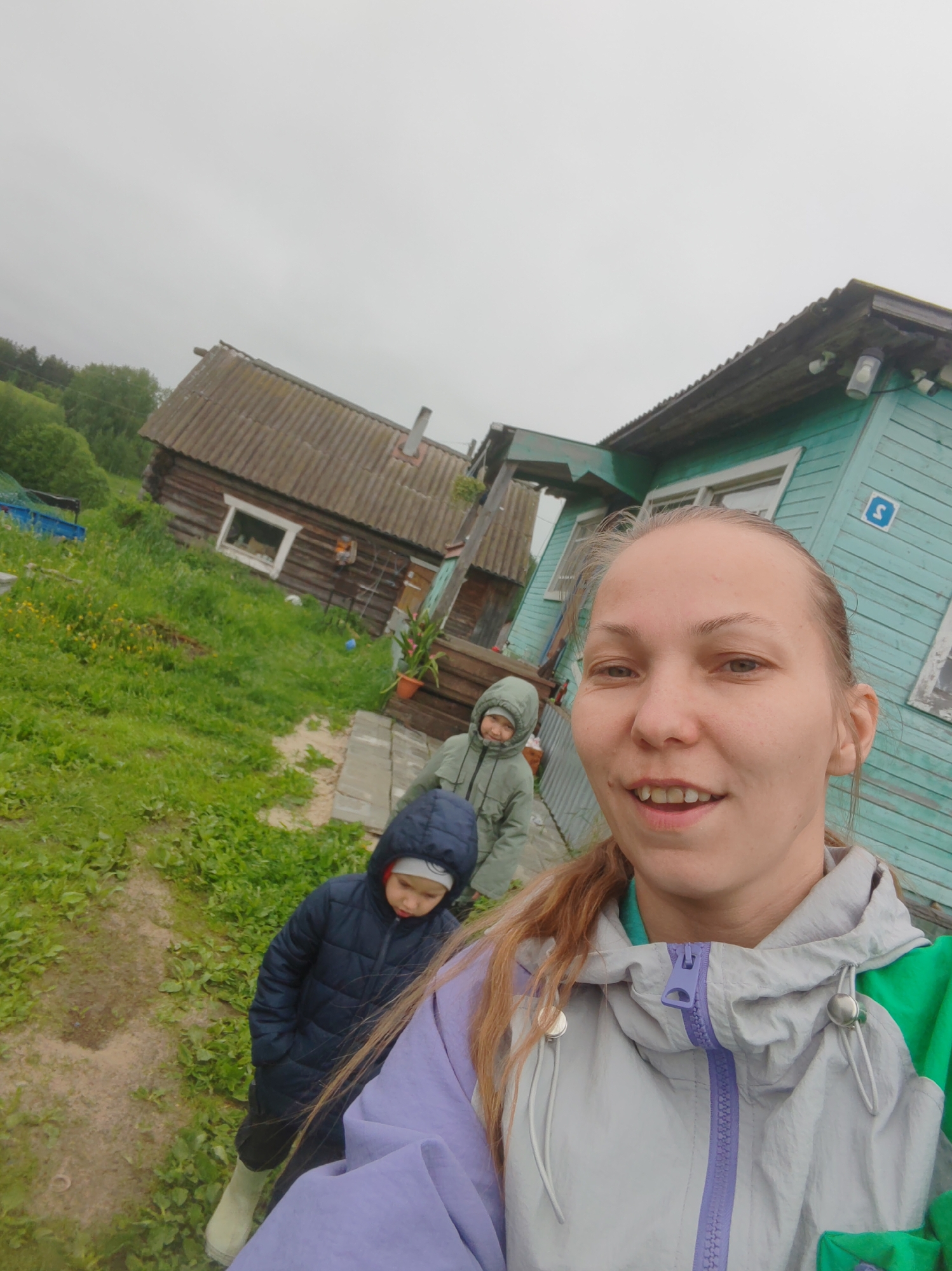 The image is a daytime selfie taken in Чухлэм, Russia, featuring a woman in the foreground and two children, along with two rural houses, in the background under an overcast sky.
The main subject is a woman, likely the photographer, in the foreground, seen from the chest up. She has light brown hair pulled back and a faint, pleasant smile on her face, revealing her teeth slightly. She is wearing a multi-colored jacket that is light grey, purple, and green, with a prominent purple zipper.
Behind her, to the left, are two children. The closer child is wearing a dark blue puffer jacket with a hood and light-colored boots, looking down. Further back and slightly to the right, another child is visible in a light green or grey hooded jacket, also looking downwards.
The setting appears to be a rural or village area. To the left of the frame, there is a traditional wooden log cabin with a corrugated roof and a chimney, featuring a single window. In front of this cabin, there is an expanse of green grass with some weeds and wildflowers, and a dirt path. To the right, another house, painted a light blue or teal, with white window frames and a similar corrugated roof, is visible. A blue square sign with the white number '3' is affixed to its exterior. A potted plant sits near the entrance of this blue house. The sky is uniformly grey and cloudy, suggesting overcast or potentially rainy weather, which aligns with the jackets worn by all individuals. The overall impression is a casual outdoor moment in a rustic environment.