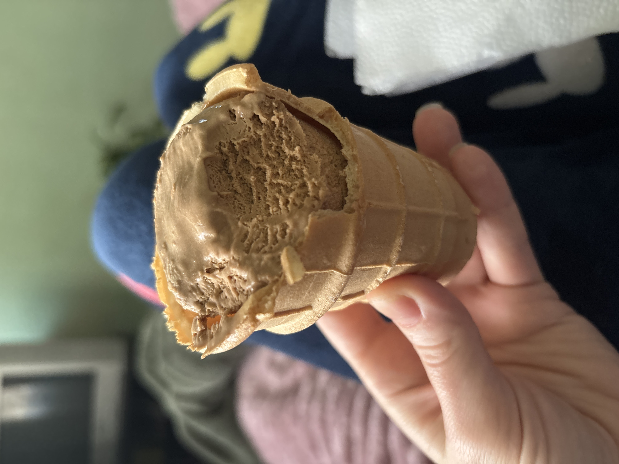 A hand holds a partially eaten, light-brown ice cream cone against a backdrop of clothing and fabric in various colors. The ice cream has a textured, slightly melted appearance. A white tissue is visible in the background. The location is Unknown, Unknown.