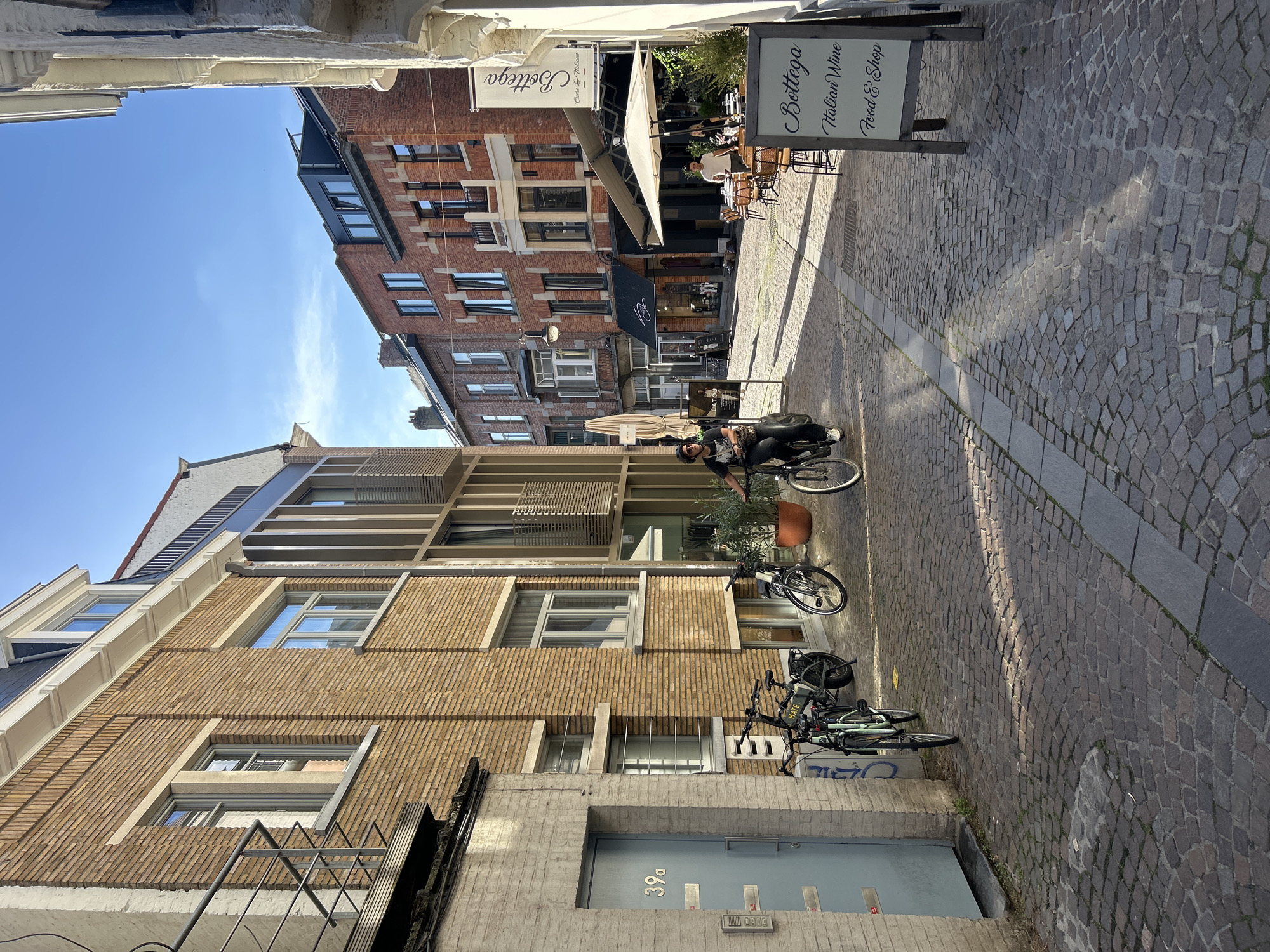 The image depicts a narrow, cobblestone street in the historic city of Leuven, Belgium, bathed in bright sunlight. On the left, a brick building with a light blue door marked "39a" stands prominently. Several bicycles are parked in front of this building. Further down the street, a person is riding a bicycle, appearing to be heading towards the viewer. On the right side of the street, a restaurant with outdoor seating is visible, adorned with umbrellas. A sign for "Bottega" advertising "Italian Wine, Food & Shop" is positioned in the foreground. The architecture suggests a European city with a mix of older brick buildings and more modern architectural elements. The sky is clear blue.