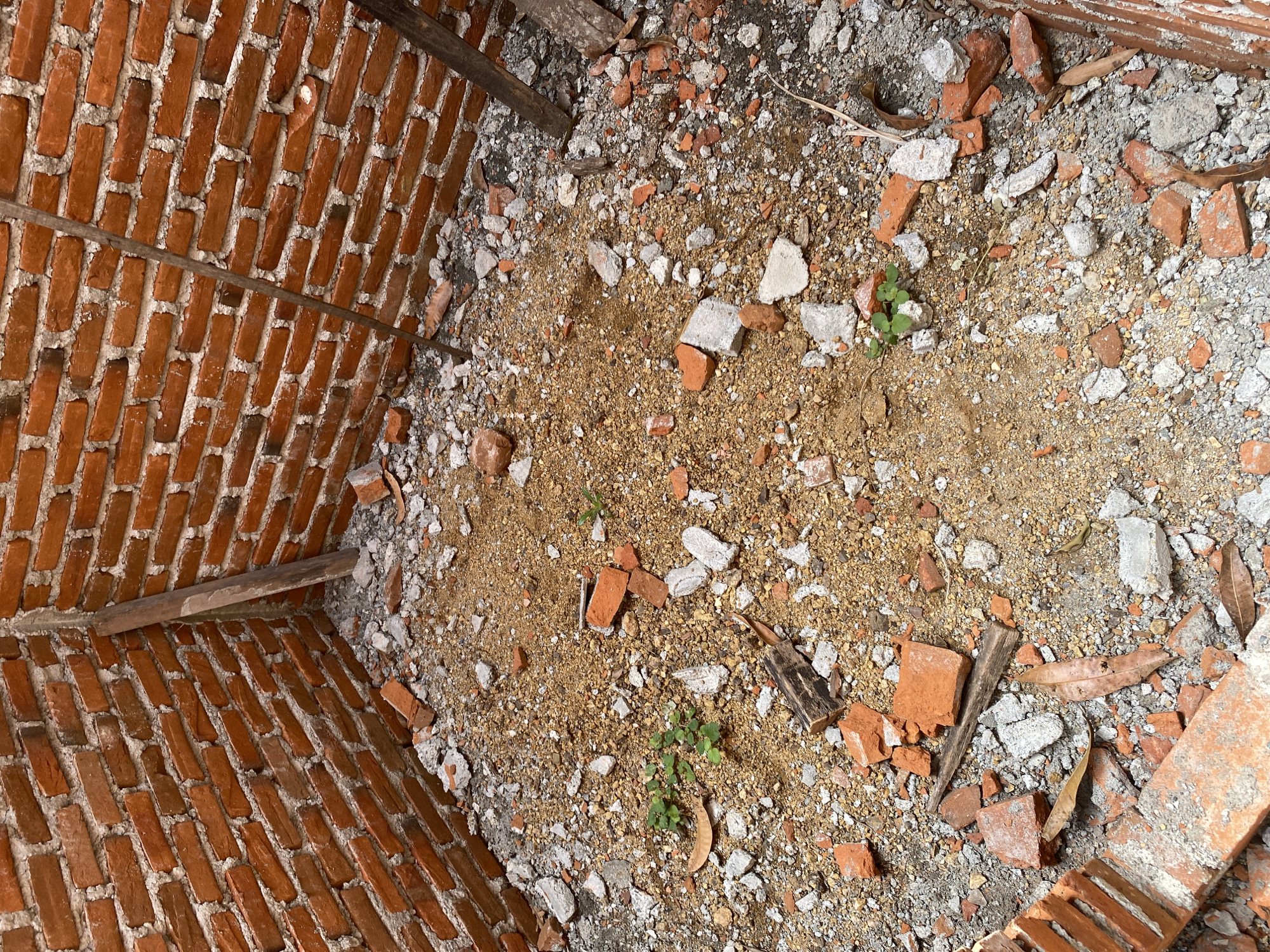 The image shows the interior of a brick building under construction, with the walls made of orange bricks and white mortar. The floor is covered with dirt, sand, and debris, including broken bricks, concrete fragments, and some small green plants. There are also several wooden planks leaning against the walls, likely used as support during construction. The scene is desolate and appears to be an unfinished construction site. The location is identified as Unknown, Unknown.