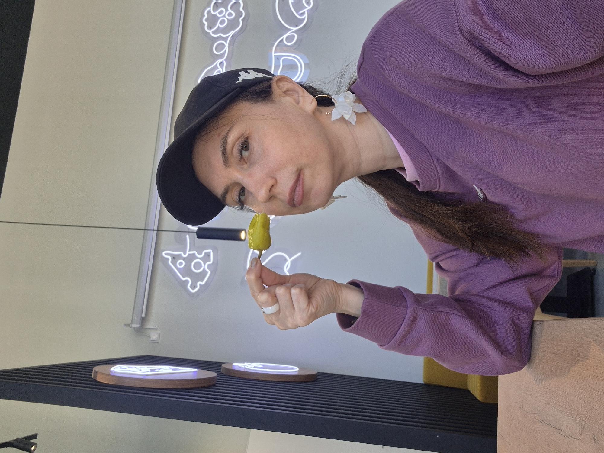 A woman with light skin, dark hair in a ponytail, and wearing a black baseball cap, white flower earrings, and a purple sweatshirt is holding a small, yellow-green pepper in her left hand near her face. She is wearing a white ring on her left ring finger. The woman is looking towards the pepper.
In the background, there is a wall with neon light designs of what appears to be food items, and a black slatted shelf with round, illuminated disks. The scene is inside, and likely in a commercial space. Based on visual details, this appears to be at the Izhevsk, Russia, pizzeria "Pizzburg."