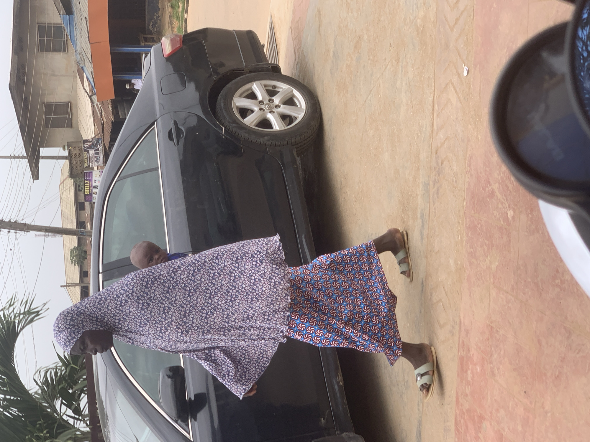 A woman wearing a blue and white floral hijab and a blue and red patterned skirt walks on a paved street next to a black sedan. A young child, visible through the car window, is being carried on the woman's back. The child is wearing a blue garment and is looking out of the car window. The woman is wearing light green sandals. Buildings with corrugated metal roofs are visible in the background. The image was taken in Shagamu, Nigeria.