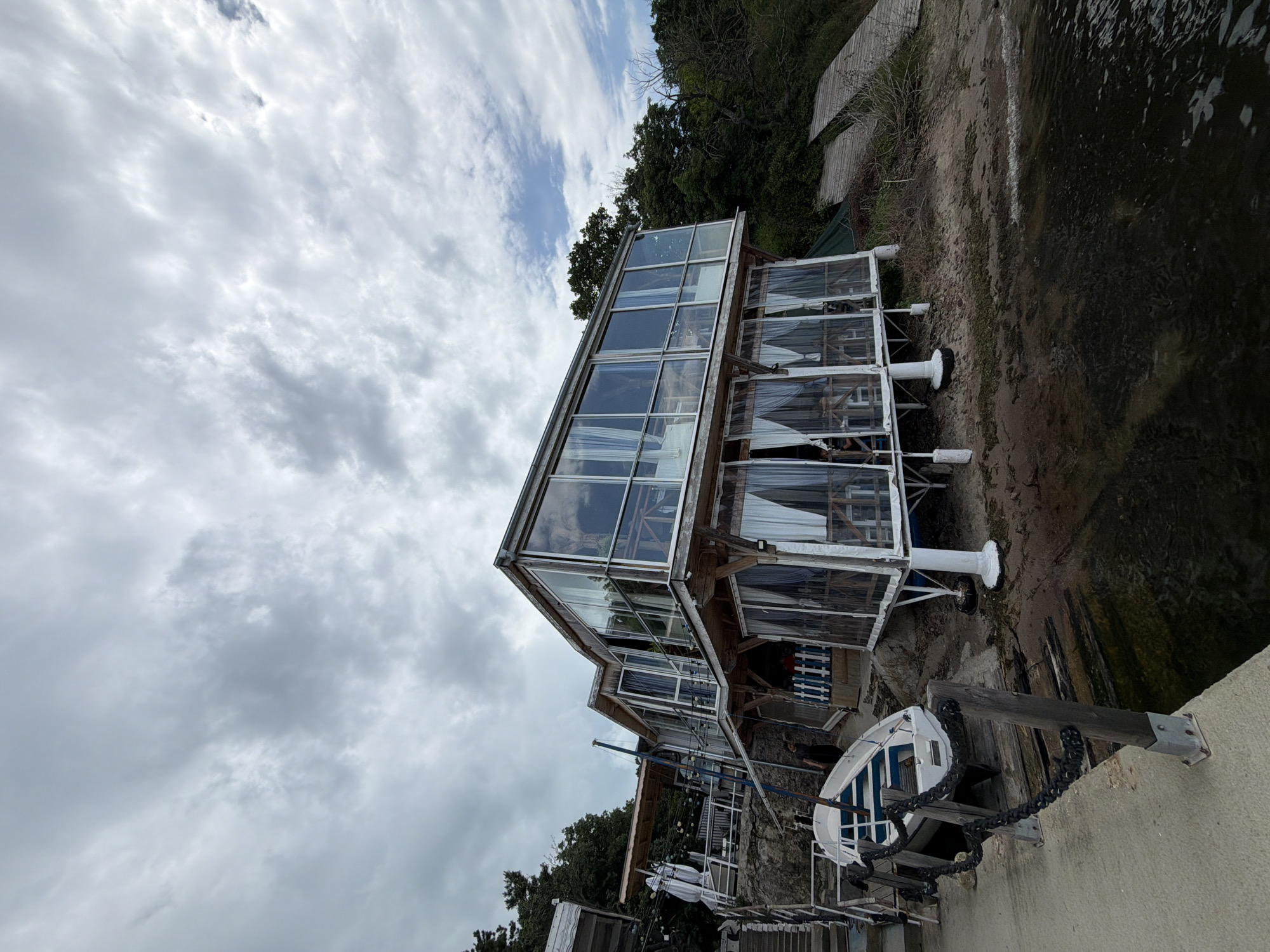 The image depicts a two-story building with a glass-enclosed structure on the water's edge under a cloudy sky. The building appears to be constructed with a wooden frame and large glass panels. It is supported by white cylindrical pillars that rise from the water or shoreline. The surrounding environment includes trees and vegetation on the hill behind the building and water bordering the building. A small boat with a blue and white hull is visible near the foreground, secured with chains to a wooden post. The scene is likely located in Burgas, Bulgaria, based on the style of architecture and the coastal environment. There are no people directly visible in the image.