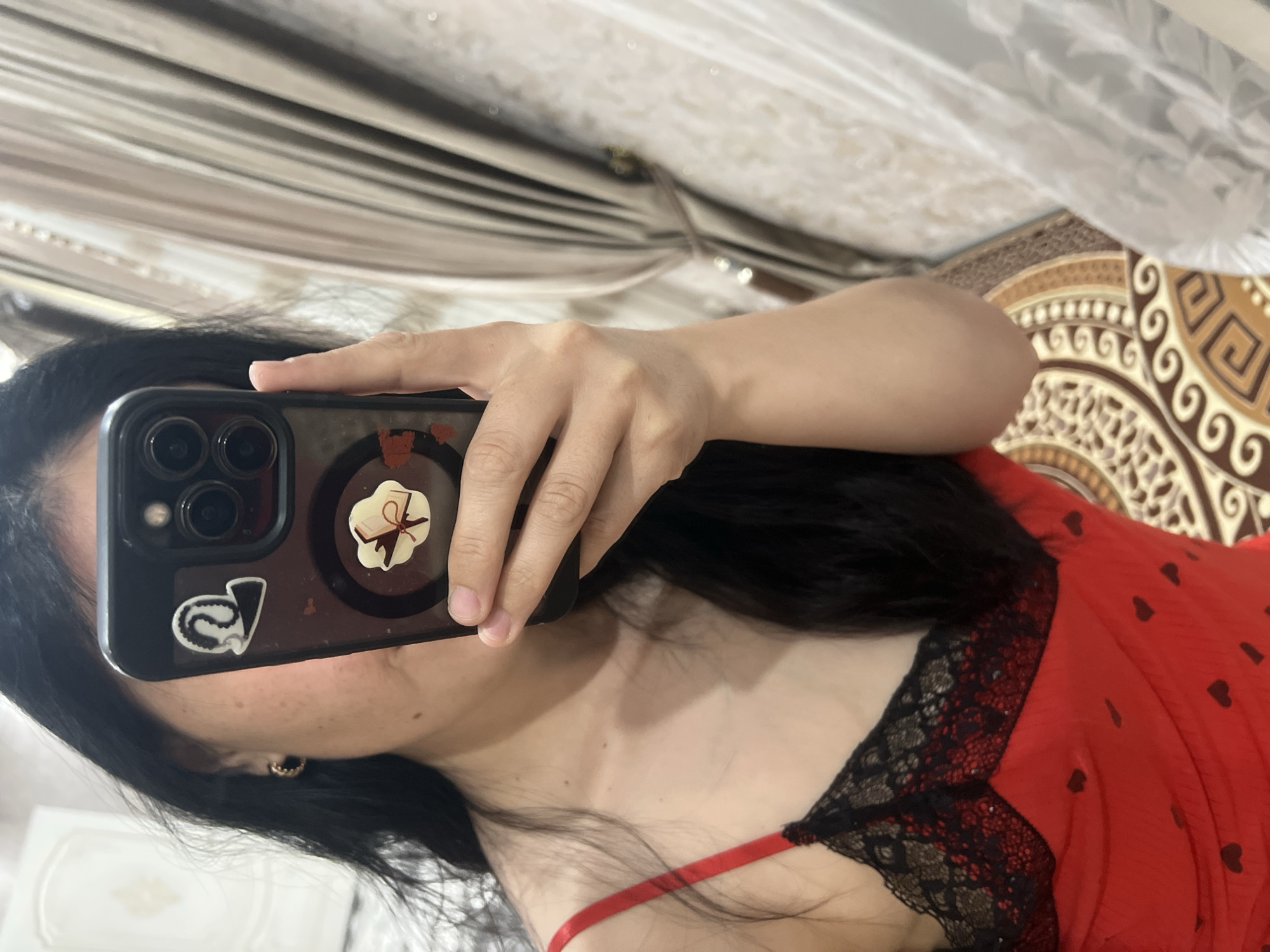 A woman is taking a selfie with her phone. She has long, dark hair and is wearing a red top with black lace trim and small dark heart patterns. The phone is held in her right hand, partially obscuring her face. The phone case has at least two stickers: one of an open book with a ribbon, and another that appears to be a stylized black letter 'V' with a chain-like design. A portion of her left ear is visible, with a small earring. The background shows a patterned rug with earthy tones and circular motifs, and a window with light-colored blinds or curtains. The overall lighting suggests an indoor setting, possibly during the daytime. The image is oriented vertically.
