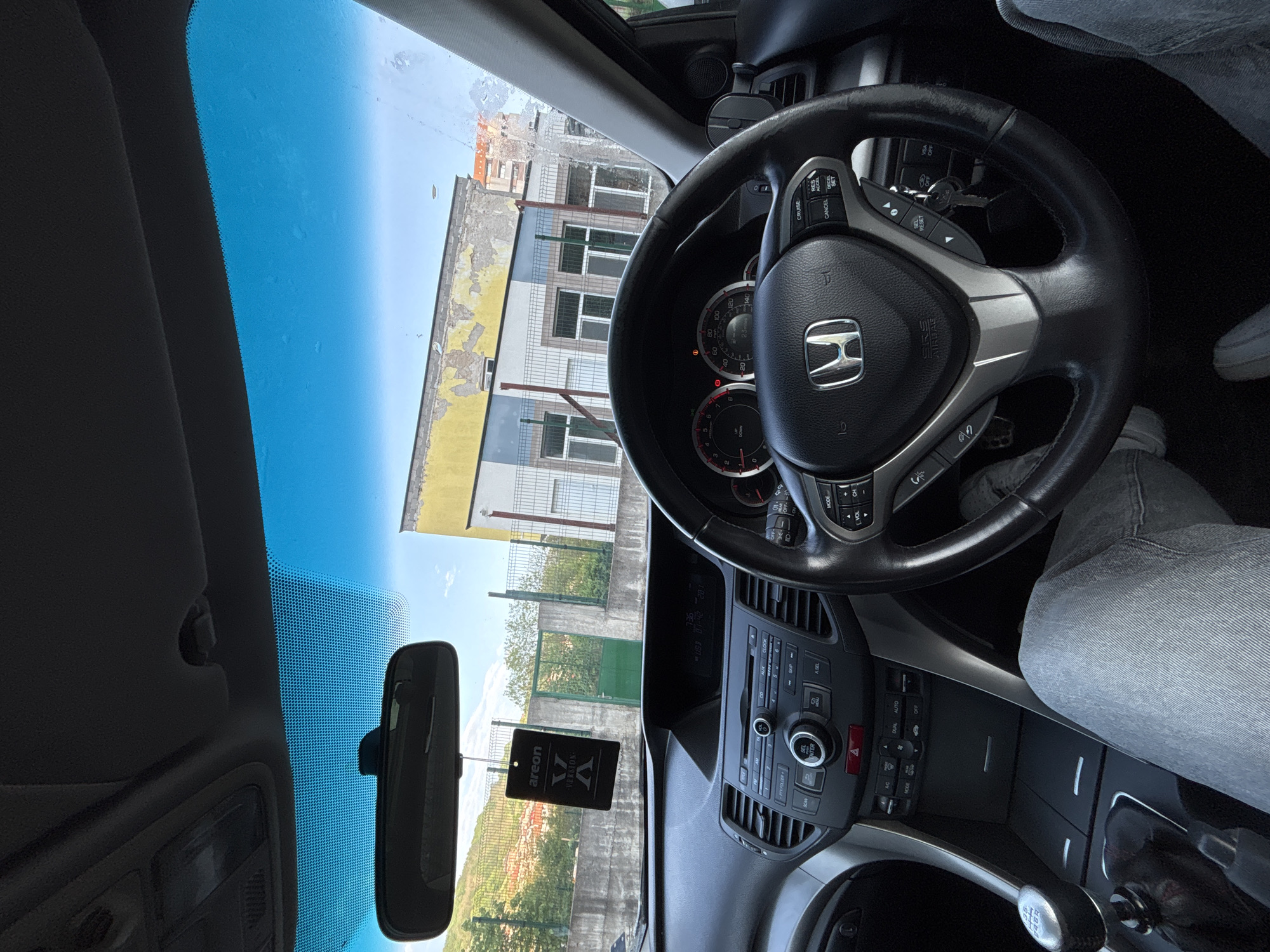 The photograph, taken from inside a car in Zlatograd, Bulgaria, captures the interior of a Honda vehicle from the driver's perspective. A black Honda steering wheel dominates the foreground, with an airbag cover at its center bearing the Honda logo. Directly behind the steering wheel, the dashboard features controls for the radio, climate, and other car systems. The rear-view mirror hangs in the middle of the windshield. An air freshener that bears the word "areon", hangs from the mirror. The windshield provides a view of the outside scenery, which includes a yellow and white building with multiple windows, some green fencing, and a partial view of the landscape. The sky is mostly blue, with some clouds. A partially visible person wearing gray pants and white shoes is sitting in the driver seat.
