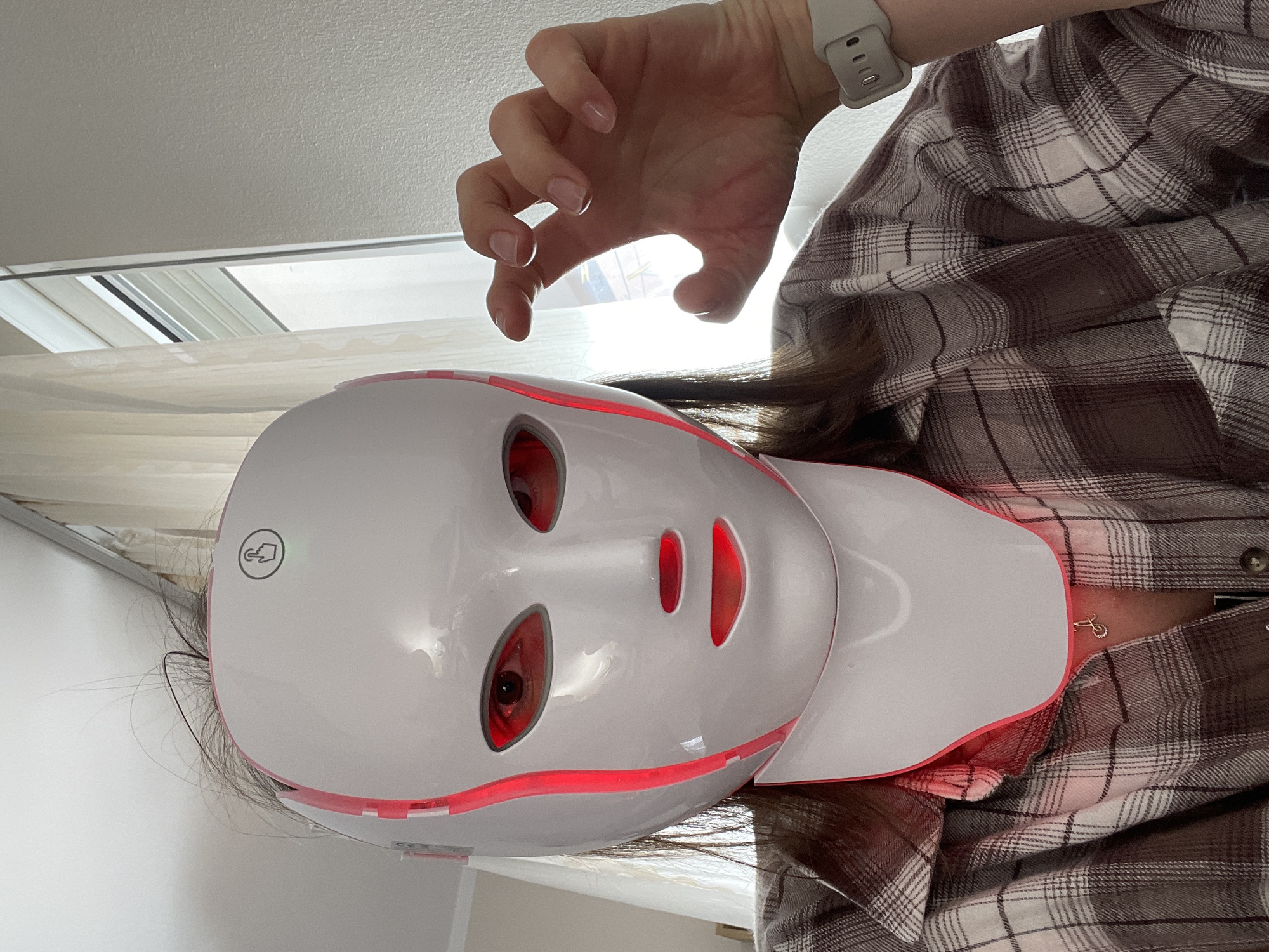 A person is wearing a white LED face mask that is lit up with red lights. The person is also wearing a plaid shirt and a watch. The background appears to be indoors, possibly in a bedroom, based on the window and wall. The person is making a claw-like gesture with their hand above the mask. There is no evidence to support the location being Budva, Montenegro.