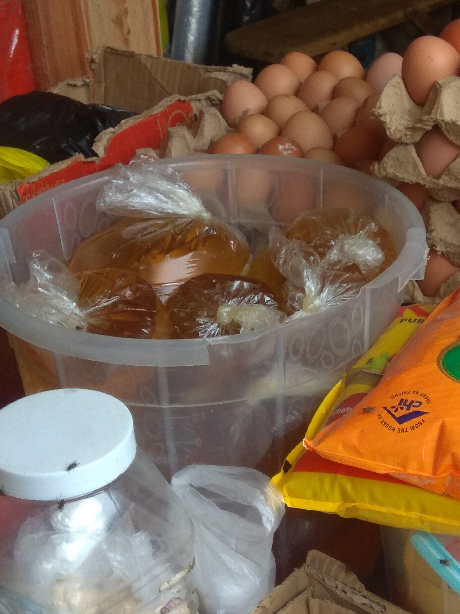 A close-up shot captures a collection of goods, likely at a market stall in Jalingo, Nigeria. The central focus is a large, clear plastic tub filled with several smaller plastic bags, each containing a yellowish-orange liquid, possibly cooking oil. These bags are sealed and appear to be a convenient way to package and sell the commodity.
Surrounding the tub are various other items. To the right, a brightly colored orange and yellow plastic bag is visible, with some text printed on it. The text, partially obscured, includes "CHI" in a blue logo and "BRAND AS FRIEND. FROM THE HOUSE OF". To the left of the tub, a transparent plastic jar with a white lid is prominent. Two flies are visible on the lid of this jar, indicating a potentially unhygienic environment.
In the background, stacks of eggs in cardboard cartons are visible, suggesting the sale of fresh produce. The scene is somewhat cluttered, with cardboard boxes and other packaging materials contributing to a busy market atmosphere. The lighting suggests it could be daytime, but the overall quality of the image, along with the presence of flies, points towards an outdoor or open-air market setting. No people are visible in the frame. The overall impression is one of everyday commerce in a Nigerian market.