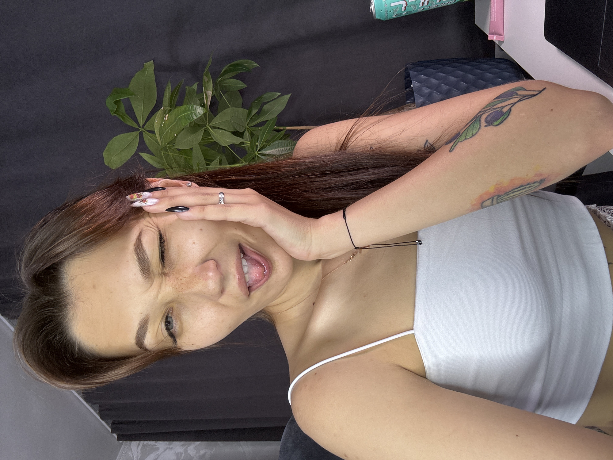 A woman with long brown hair and freckles is winking and sticking her tongue out. She is wearing a white tank top and has long acrylic nails with nail art. She has tattoos on her arm. She is holding her hand to her cheek and wearing rings on her fingers. A plant is visible in the background. The image appears to be taken indoors, possibly in an apartment or office setting. The woman's pose and expression suggest a playful or casual mood. There is no indication of the city of Novokuznetsk, Russia.