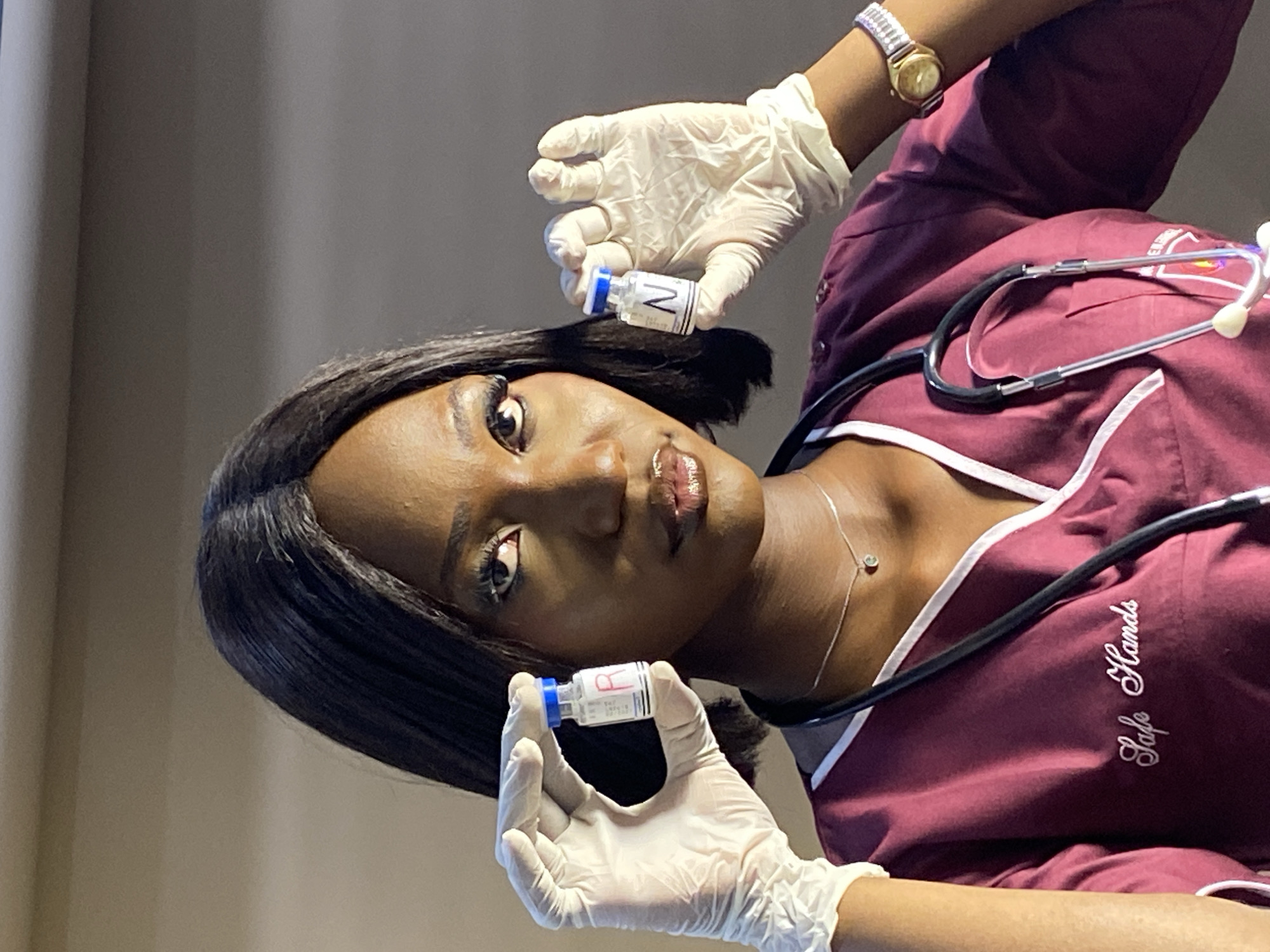 A dark-skinned woman, presumably a healthcare professional, holds two vials of medication in her gloved hands. The woman wears a maroon scrub top with white trim, a stethoscope around her neck, and a wristwatch on her left wrist. The scrub top has the phrase "Safe Hands" embroidered on it. The background is a plain, light-colored wall. The image provides no information indicating the city of Gura Top, Nigeria, so no information can be given about the location.