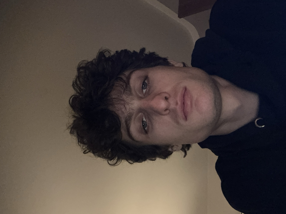 The image presents an upward-angled, head-and-shoulders portrait of a young adult male. The subject has medium-toned skin, dark, curly hair, and light-colored eyes. His expression is neutral, and he is looking directly forward, towards the camera. He wears a dark, possibly black or navy, hooded sweatshirt with a visible silver-colored metal eyelet for the drawstring located on the left side of his chest. The lighting is dim, appearing to originate from above and slightly in front of the subject. The background consists of an indistinct, light-colored plain wall. To the far right, a subtle, darker, curved or angled architectural feature is partially visible. The scene is indoors. The specific location within the city of Vincennes, France, cannot be determined from the visual information presented in the image.